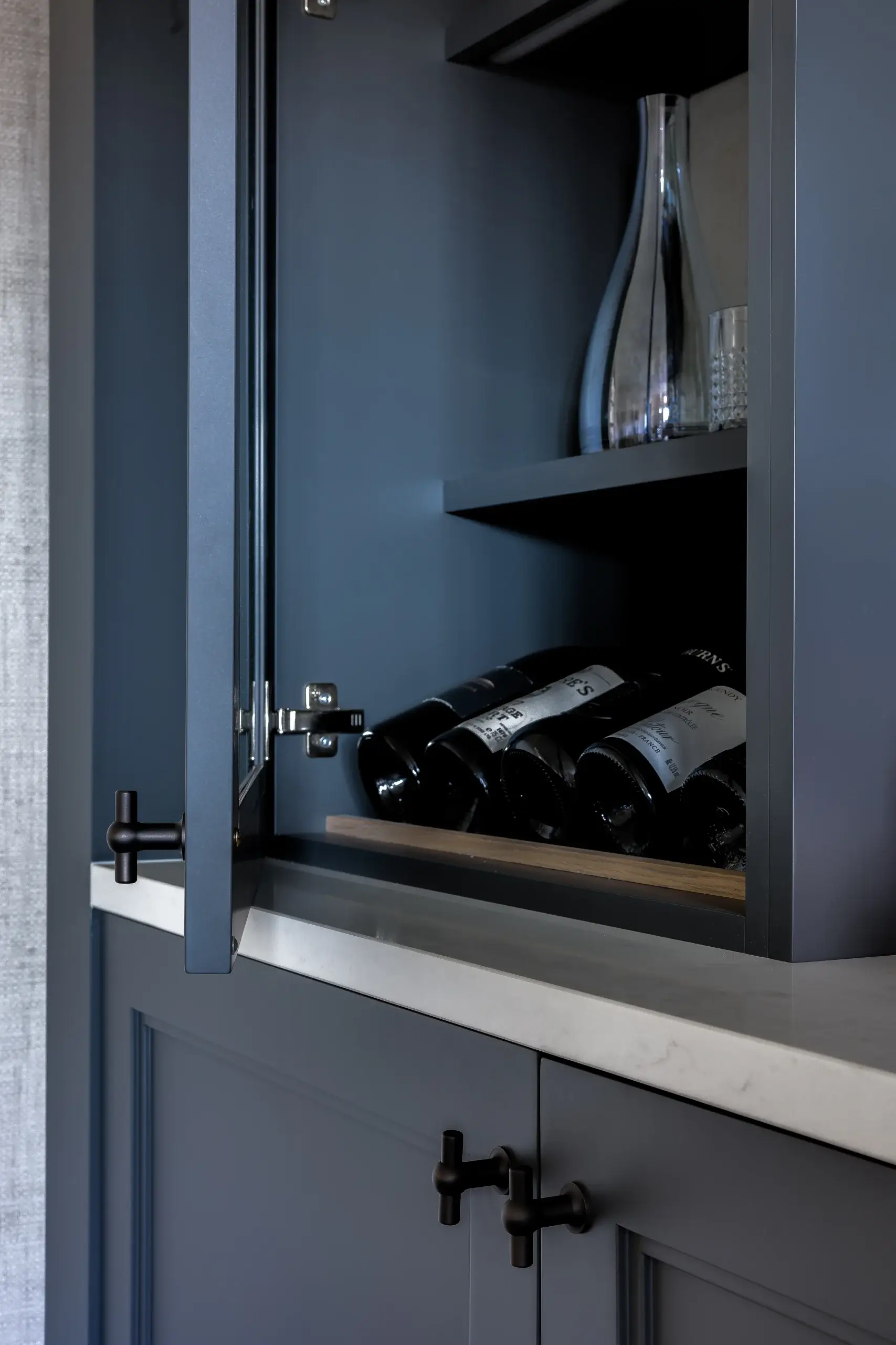 Matte navy kitchen cabinet with open glass-front door revealing wine bottles on a shelf and a tall glass carafe above.