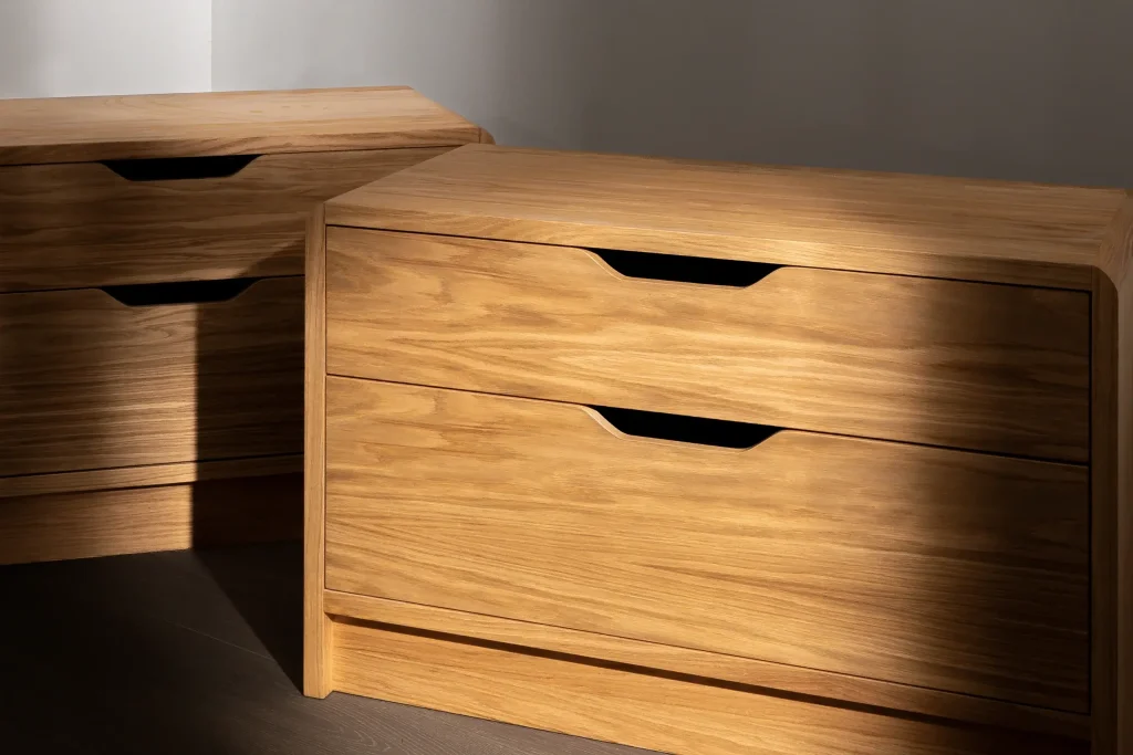 Two wooden bedside cabinets with smooth grain and recessed black finger pulls, lit by warm directional light.