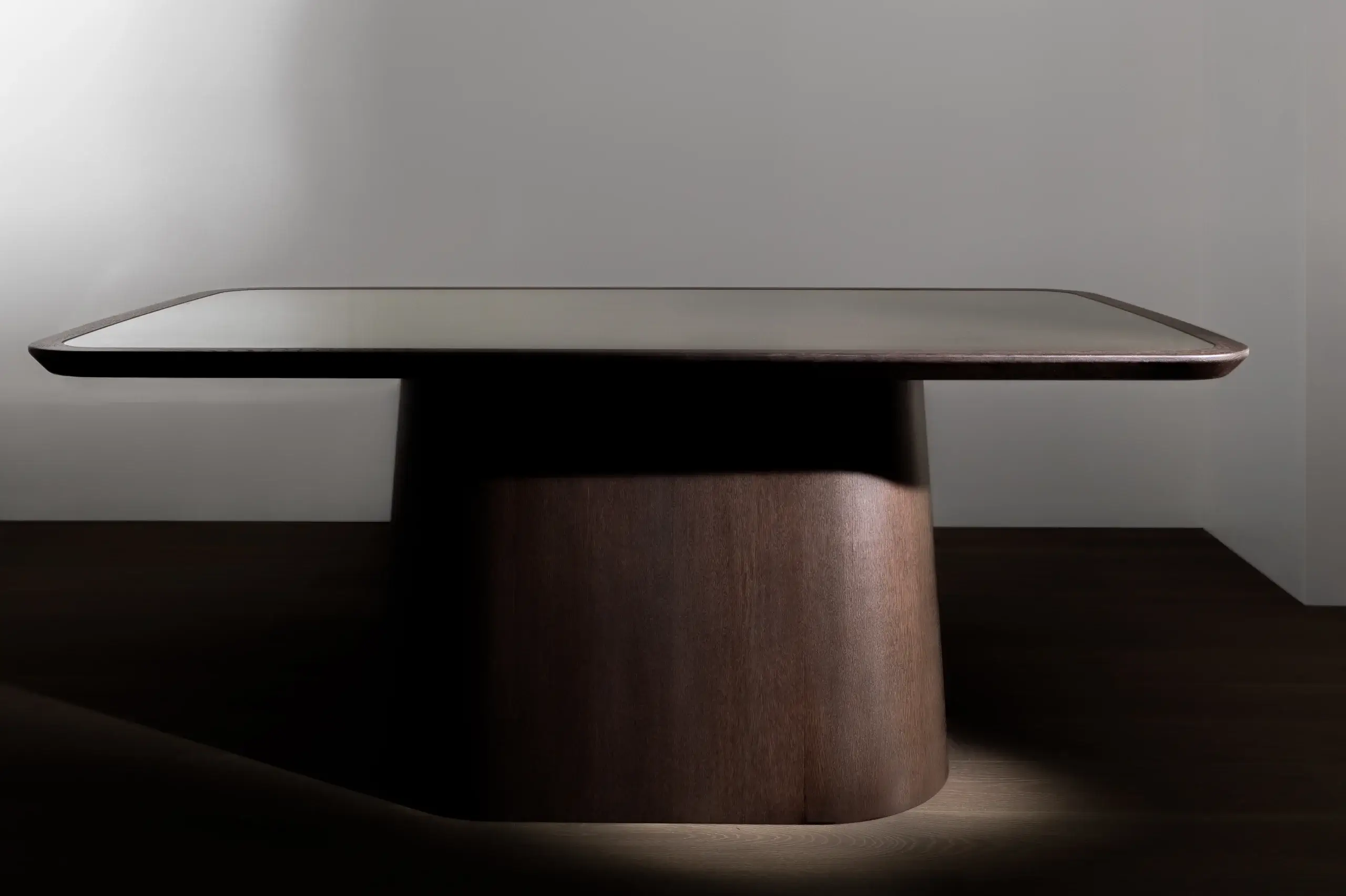 Low, modern rectangular wooden table with rounded edges and a solid cylindrical pedestal base in soft studio lighting.