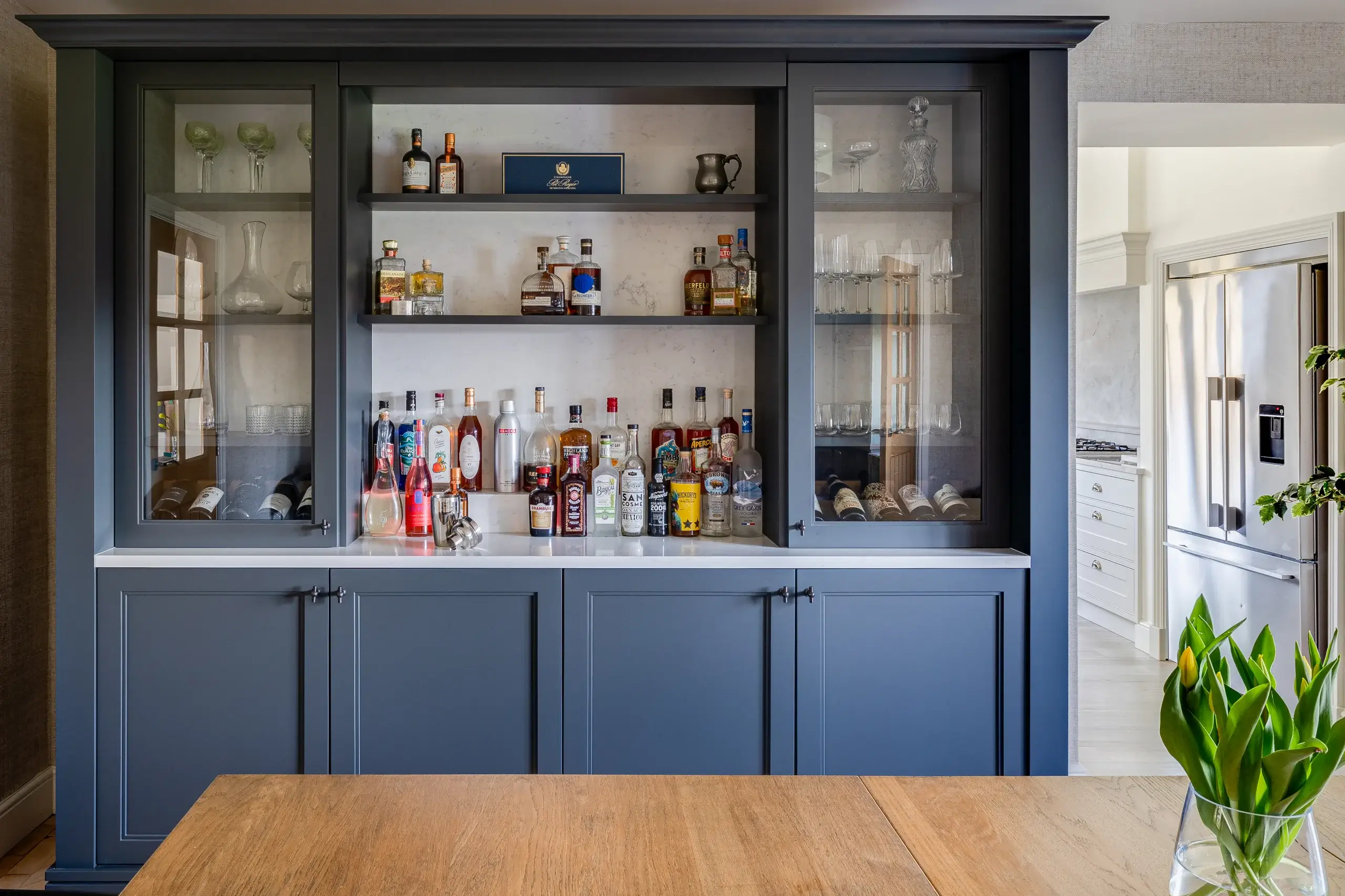 Built-in navy blue bar cabinet with glass-front shelves displaying bottles, wine, glassware, and a white countertop.