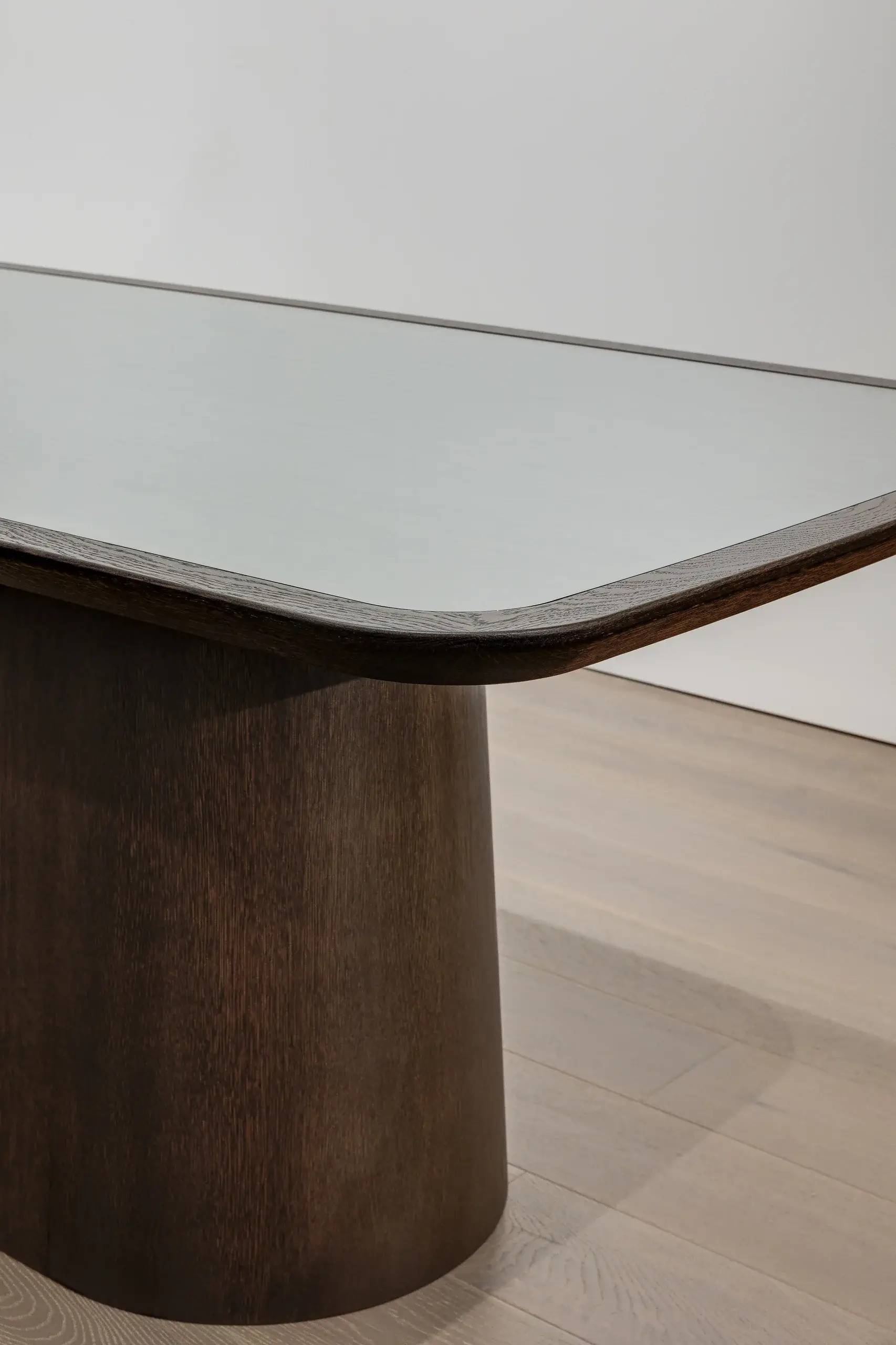 Rounded dark wood table base supporting a rectangular glass tabletop with beveled wooden edge on light hardwood floor
