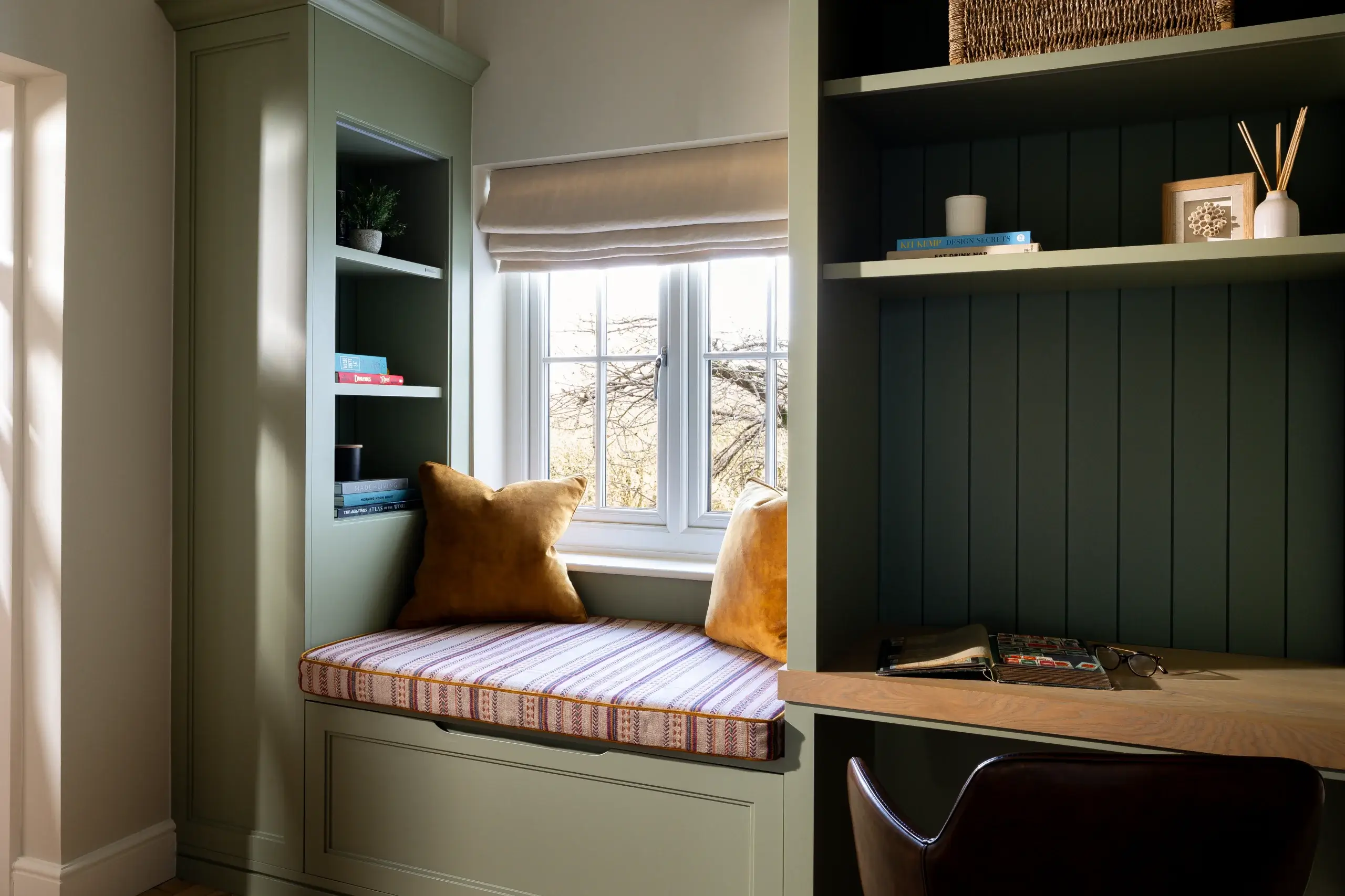 Cozy window seat with striped cushion and mustard pillows built into green painted cabinets beside a wooden desk and shelves.