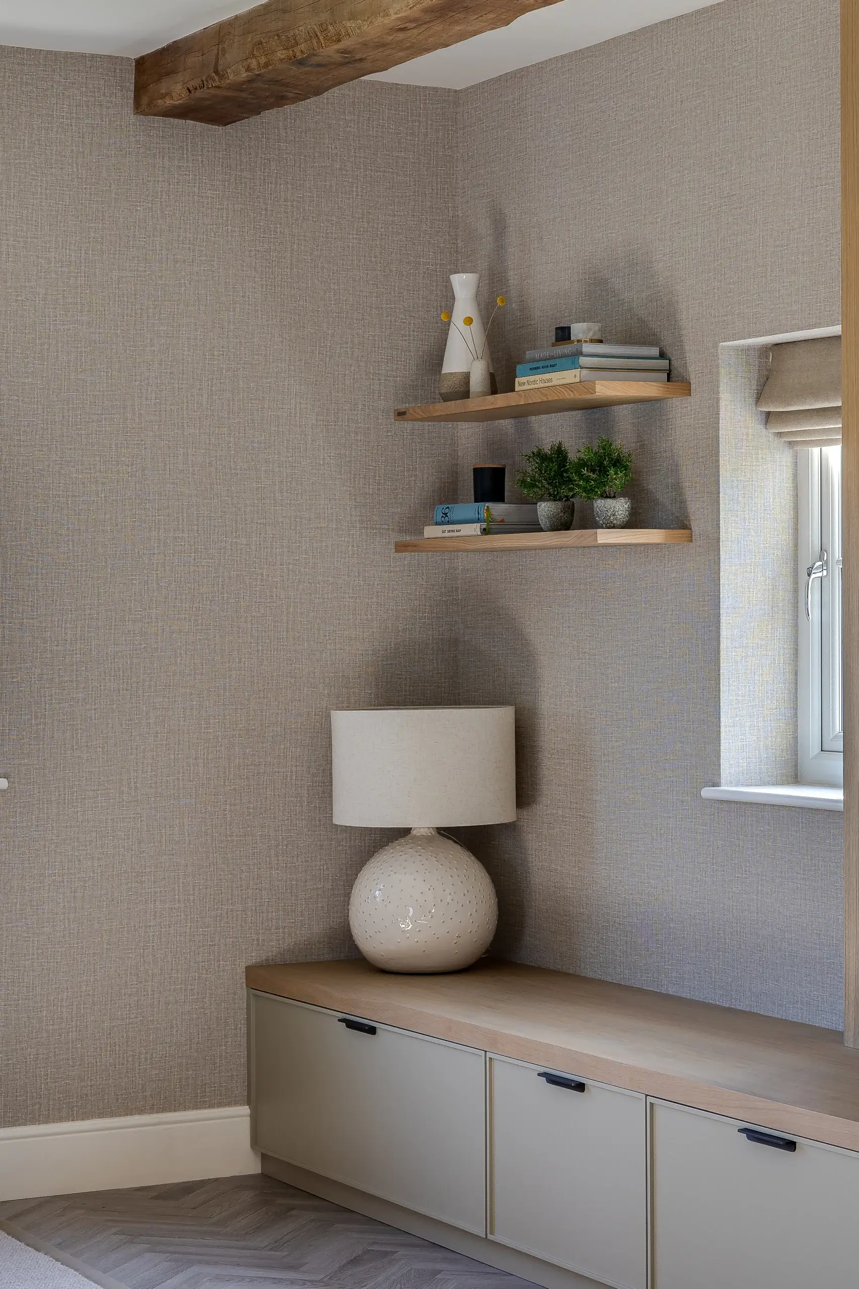 Corner of a neutral-toned room with floating wooden shelves holding books and plants, a ceramic table lamp on a low cabinet.