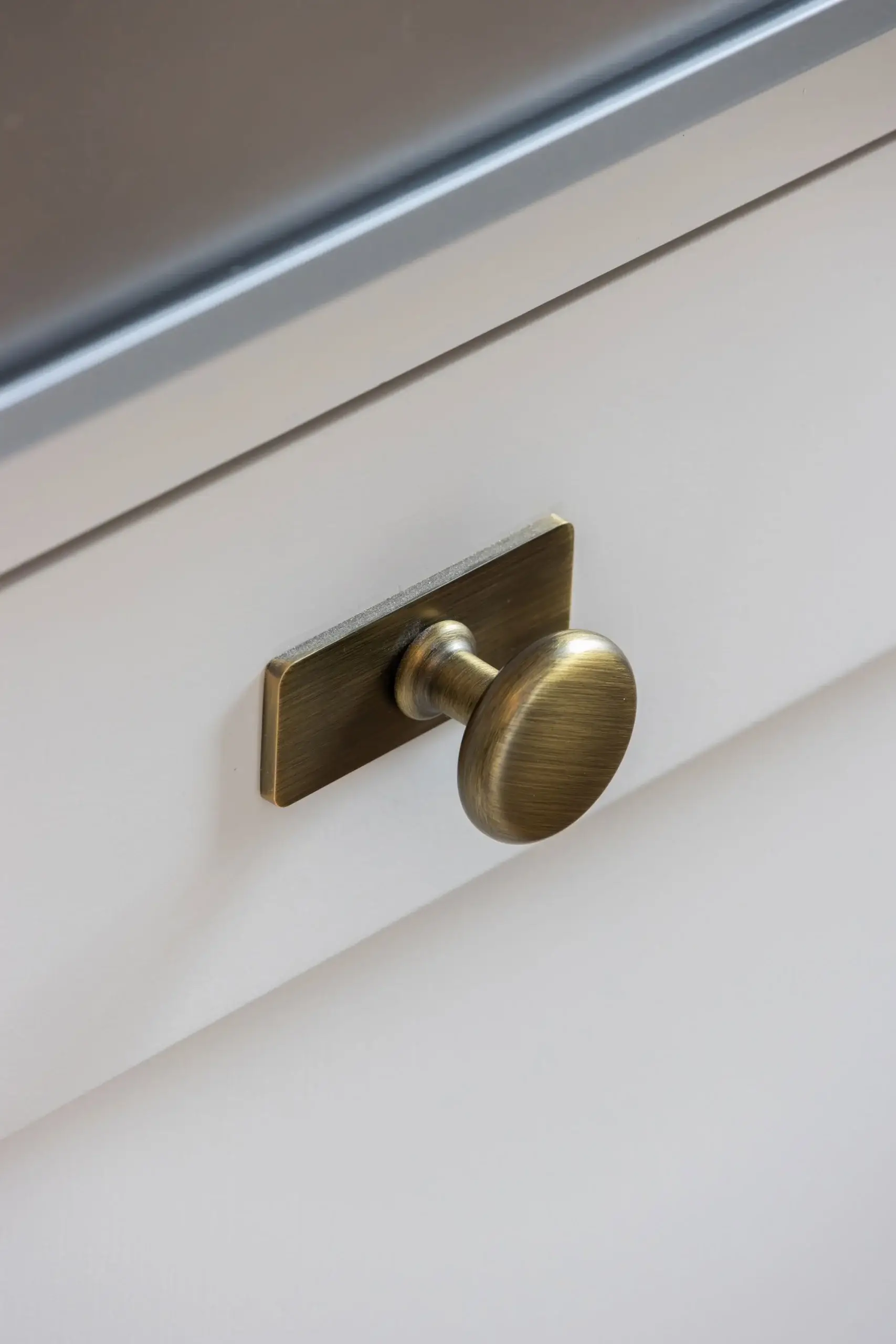 Antique brass round cabinet knob mounted on a small rectangular backplate on a white drawer front under a metal countertop