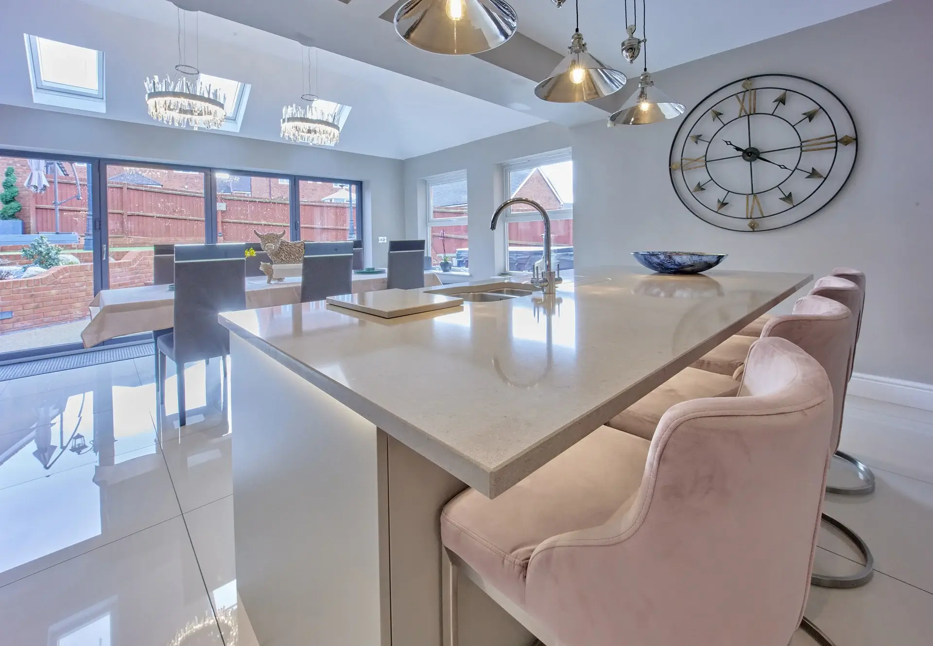 Bright modern kitchen with large island, pink velvet bar stools, chrome pendant lights, wall clock, and dining area by glass doors