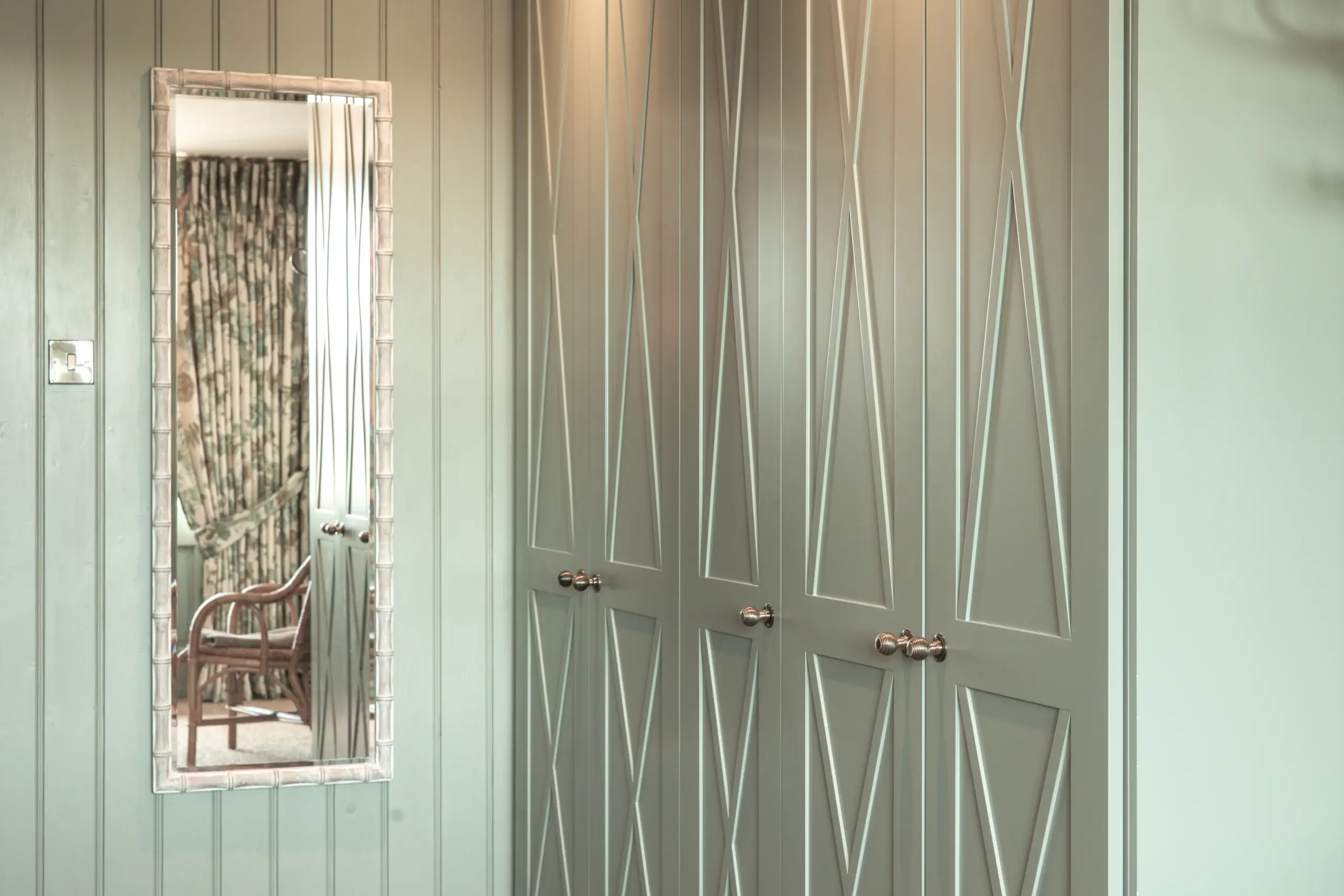 Tall rectangular wall mirror reflecting a wicker chair and floral curtains, mounted next to pale green paneled closet doors with X-patterned trim and brass knobs.