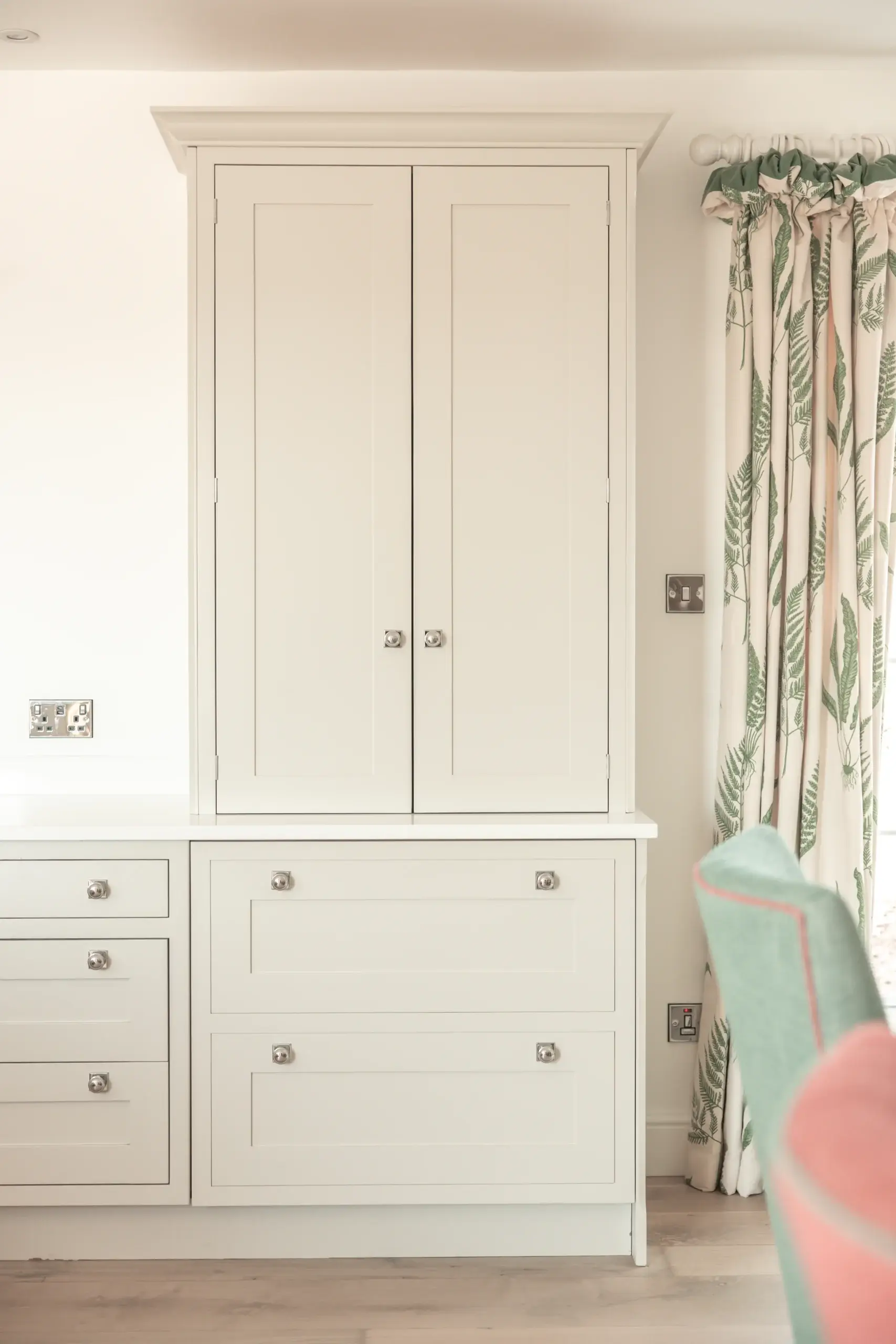 Built-in cream cabinetry with double doors above two large drawers, chrome knobs, and leafy patterned curtains at right.