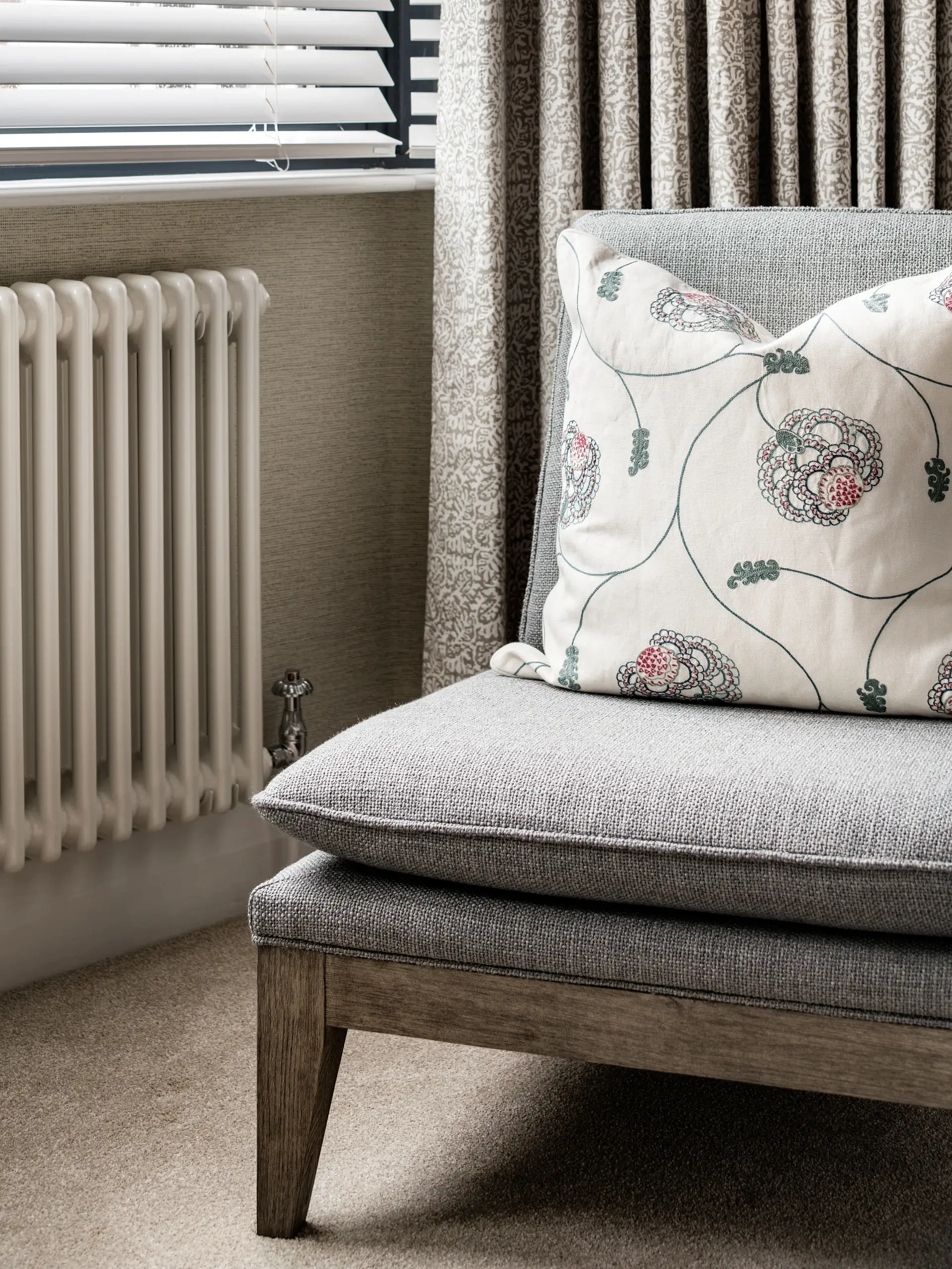Gray upholstered armchair with wooden legs and a white floral-embroidered throw pillow next to a cream radiator and patterned curtains