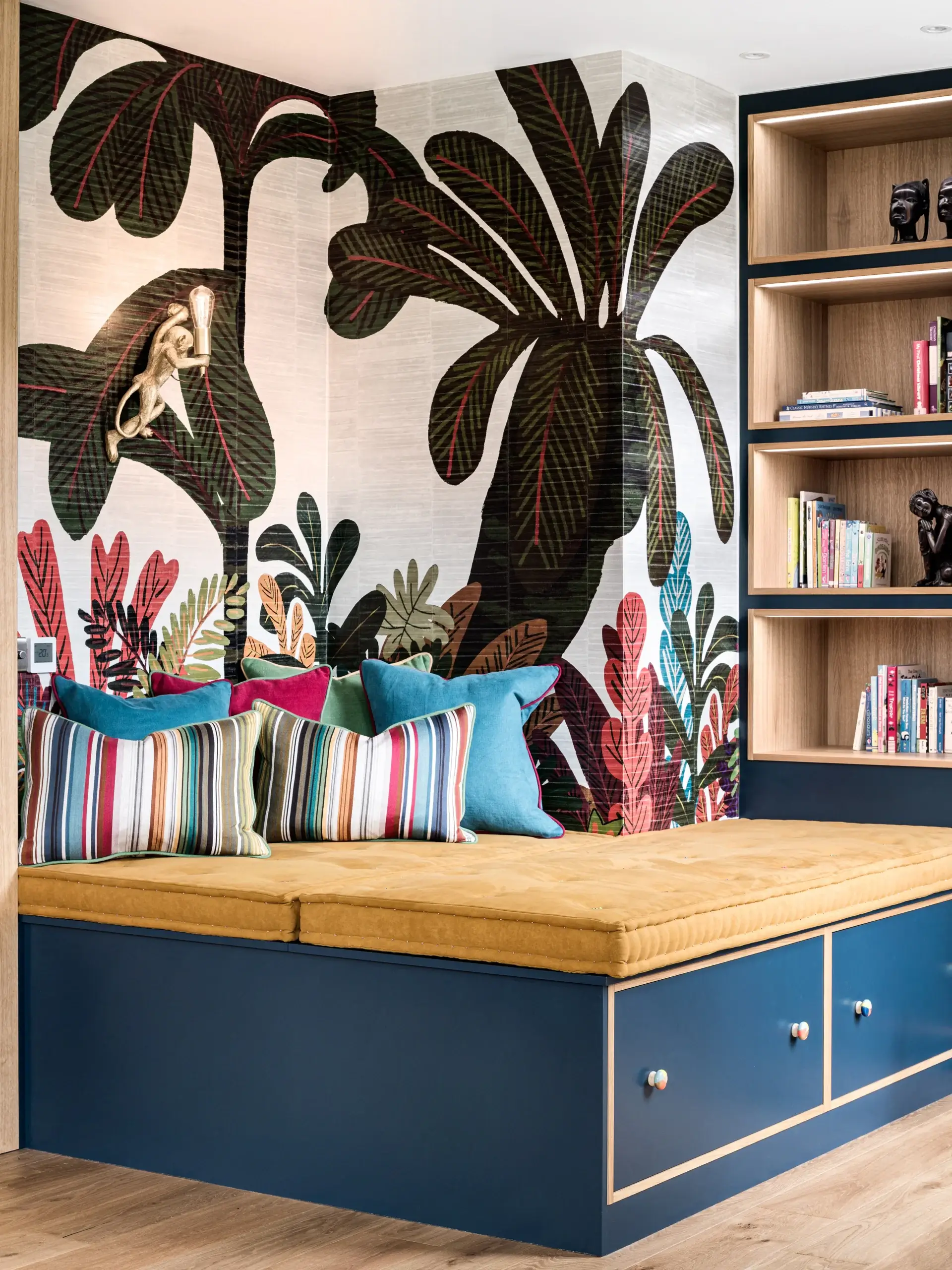 Built-in navy window seat with mustard cushion, striped and teal throw pillows, and a bold tropical leaf mural on the wall beside wooden bookshelves.