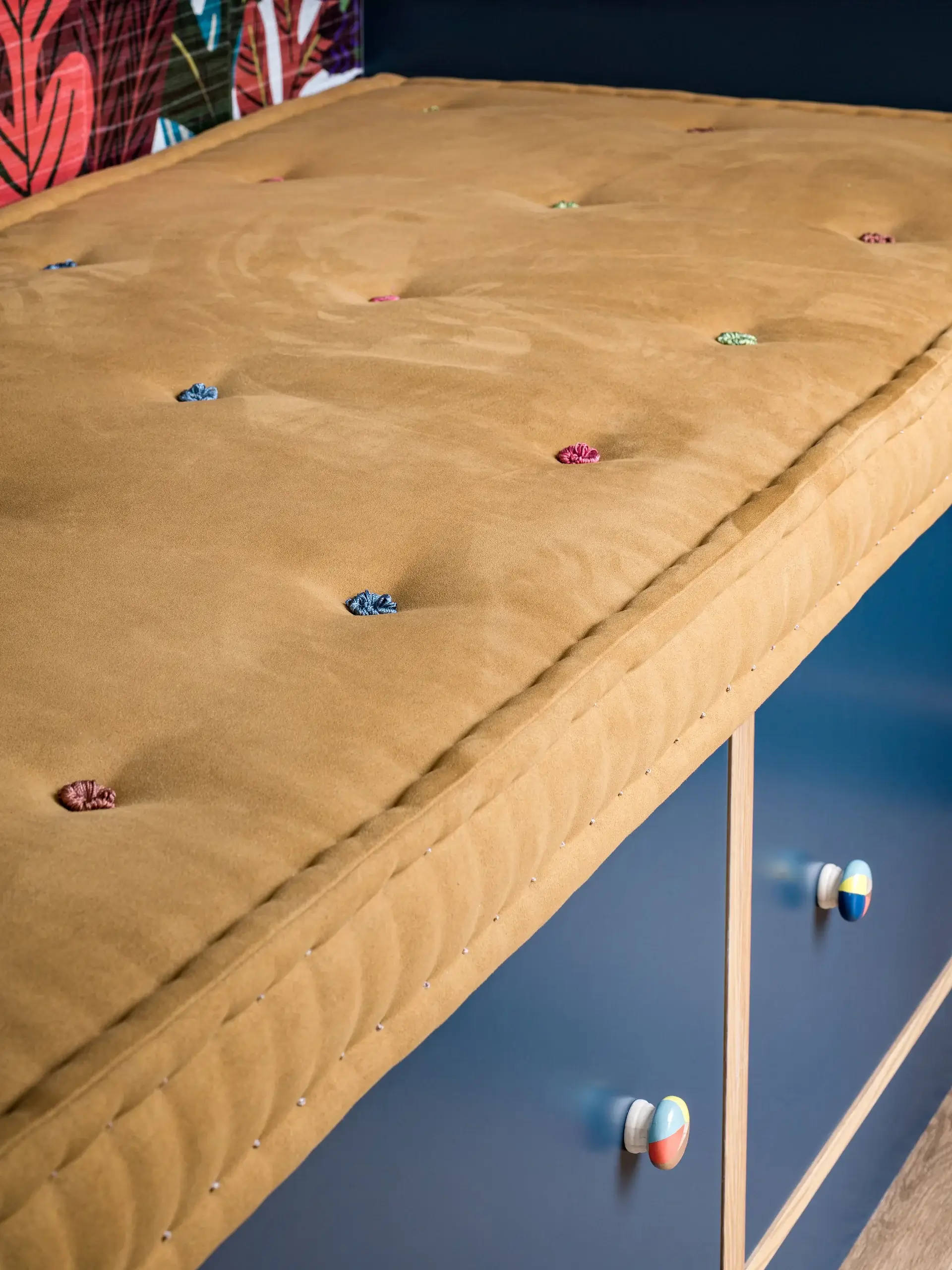 Tufted mustard-colored bench cushion with multicolored fabric buttons resting on blue wooden storage drawers.