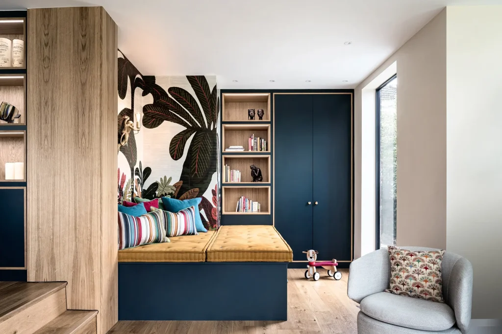 Built-in navy daybed with mustard cushion, colorful striped pillows, tropical wallpaper, bookshelves and a toy scooter by a window