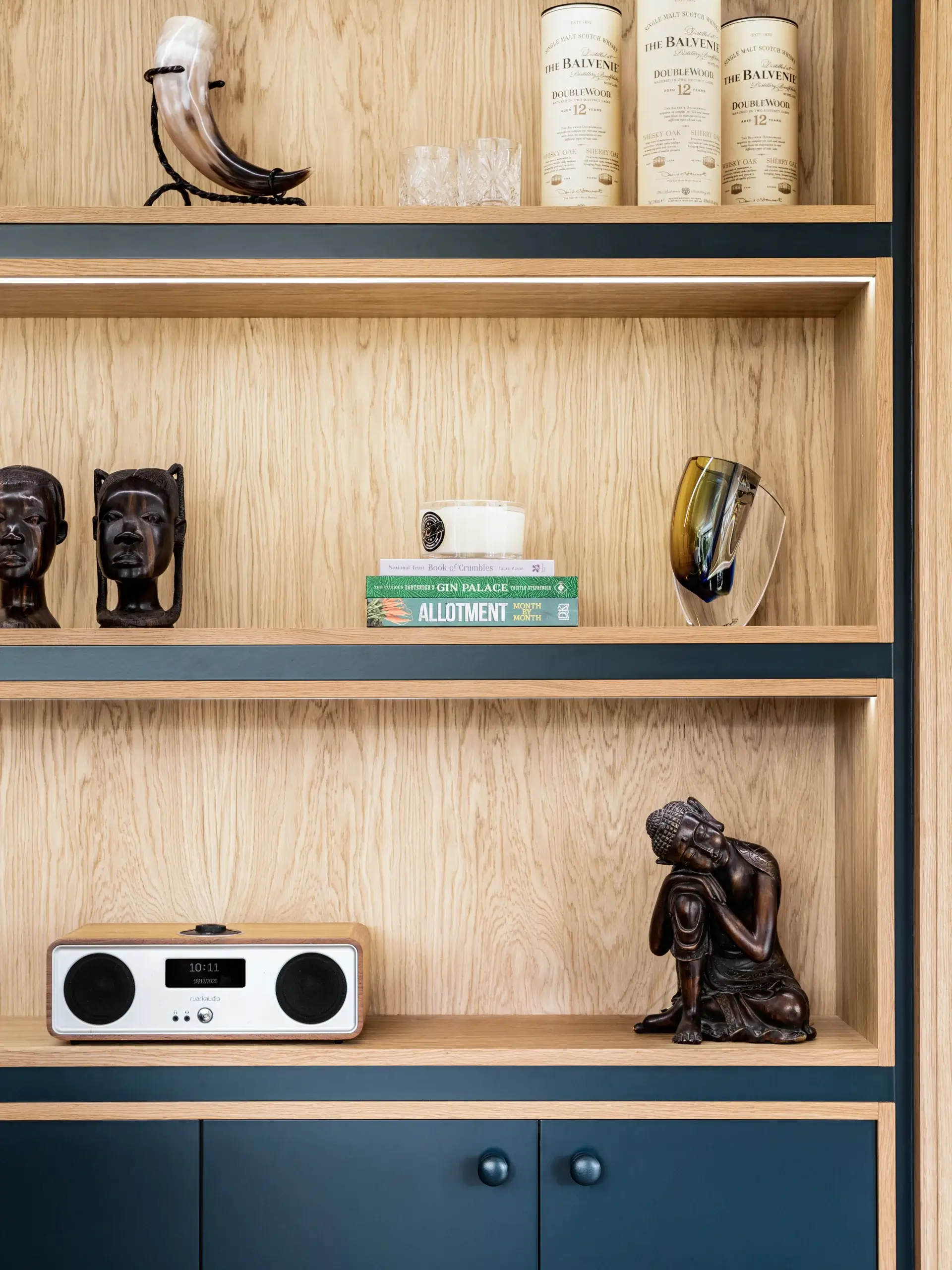 Wooden shelving unit with decorative items: carved dark wood heads, stacked books and candle, glass sculpture, vintage radio, and seated bronze Buddha statue.