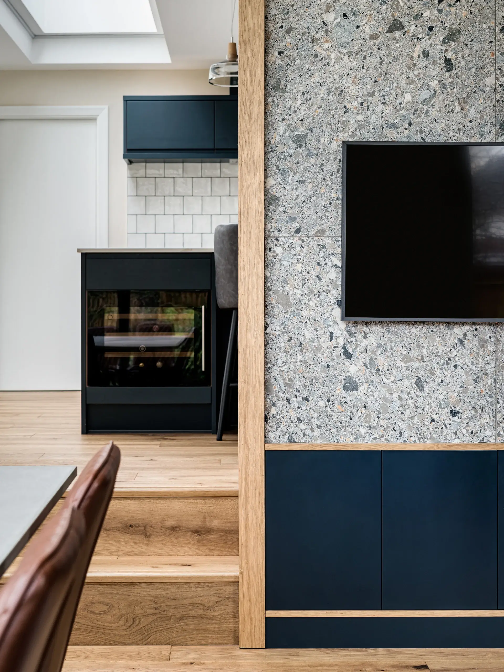 Modern interior showing oak steps, terrazzo wall with mounted TV, and navy blue cabinetry leading to a kitchen island.