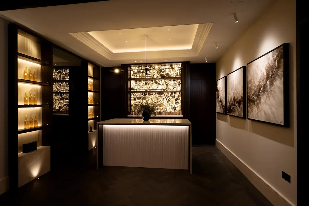 Dimly lit luxurious home bar with illuminated backlit stone wall, central island, glass bottle shelves, and abstract wall art.