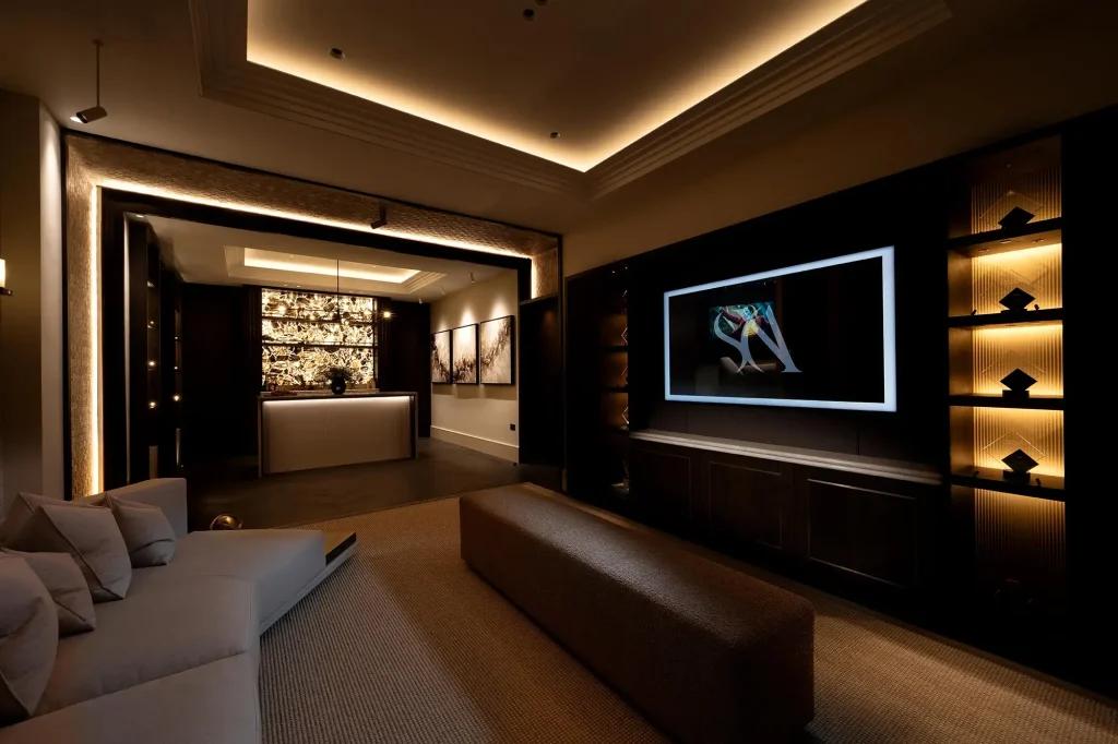 Luxurious dimly lit home theater with recessed lighting, large wall-mounted TV, plush sofa, and illuminated shelving and bar area