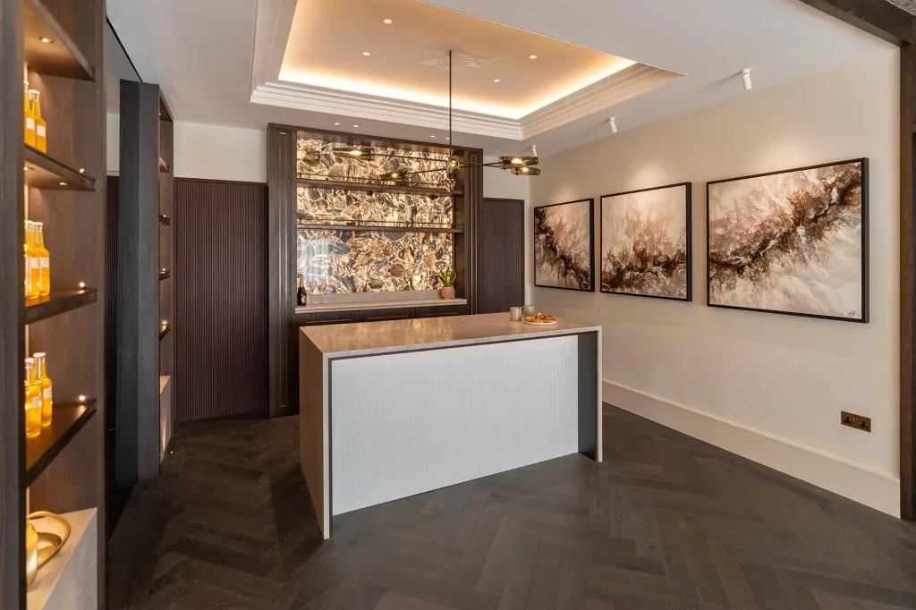 Modern kitchen island with marble countertop, backlit stone backsplash, dark herringbone floor, and three abstract wall paintings.