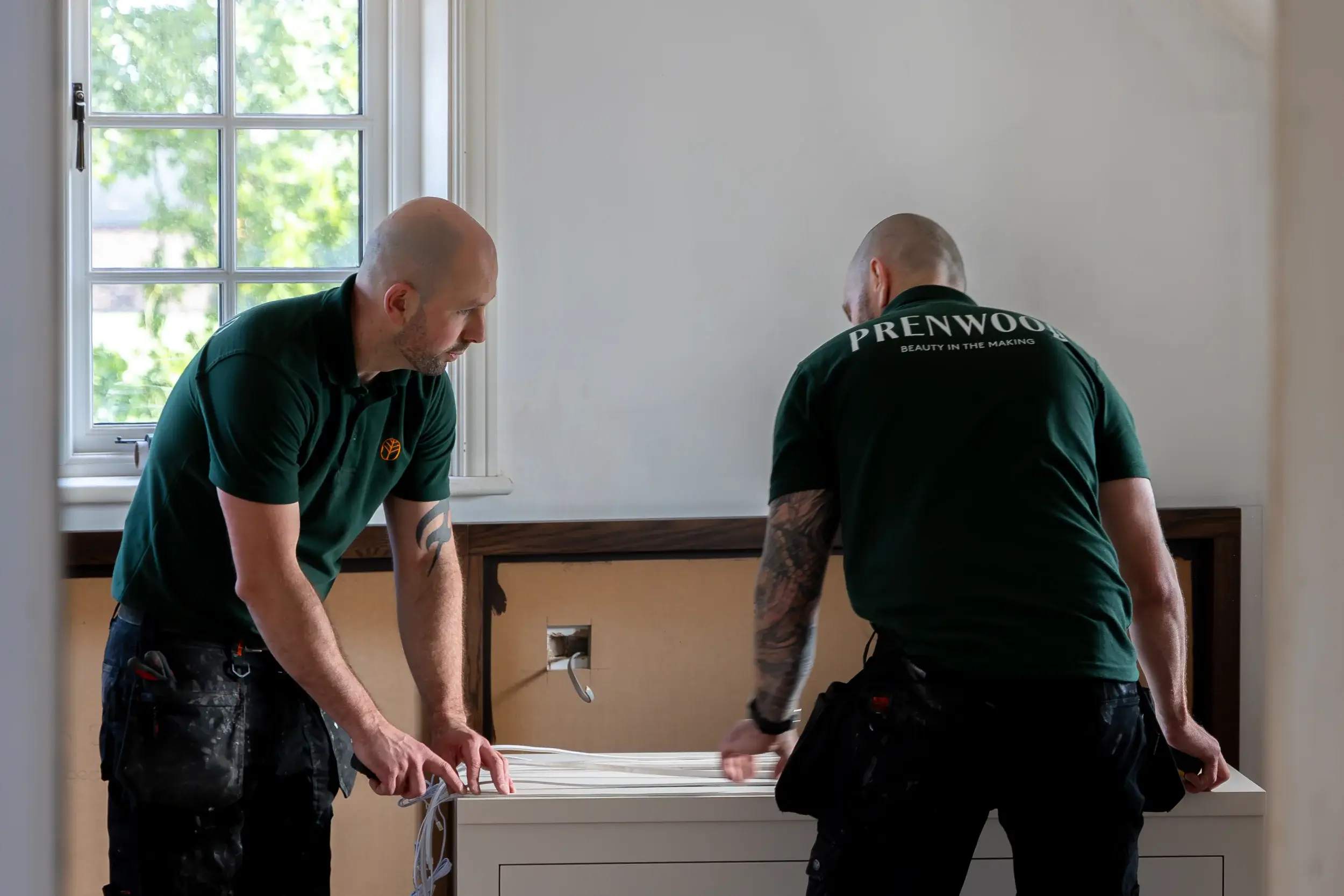 Two craftsmen in green Prenwood shirts installing cabinetry beside a window.