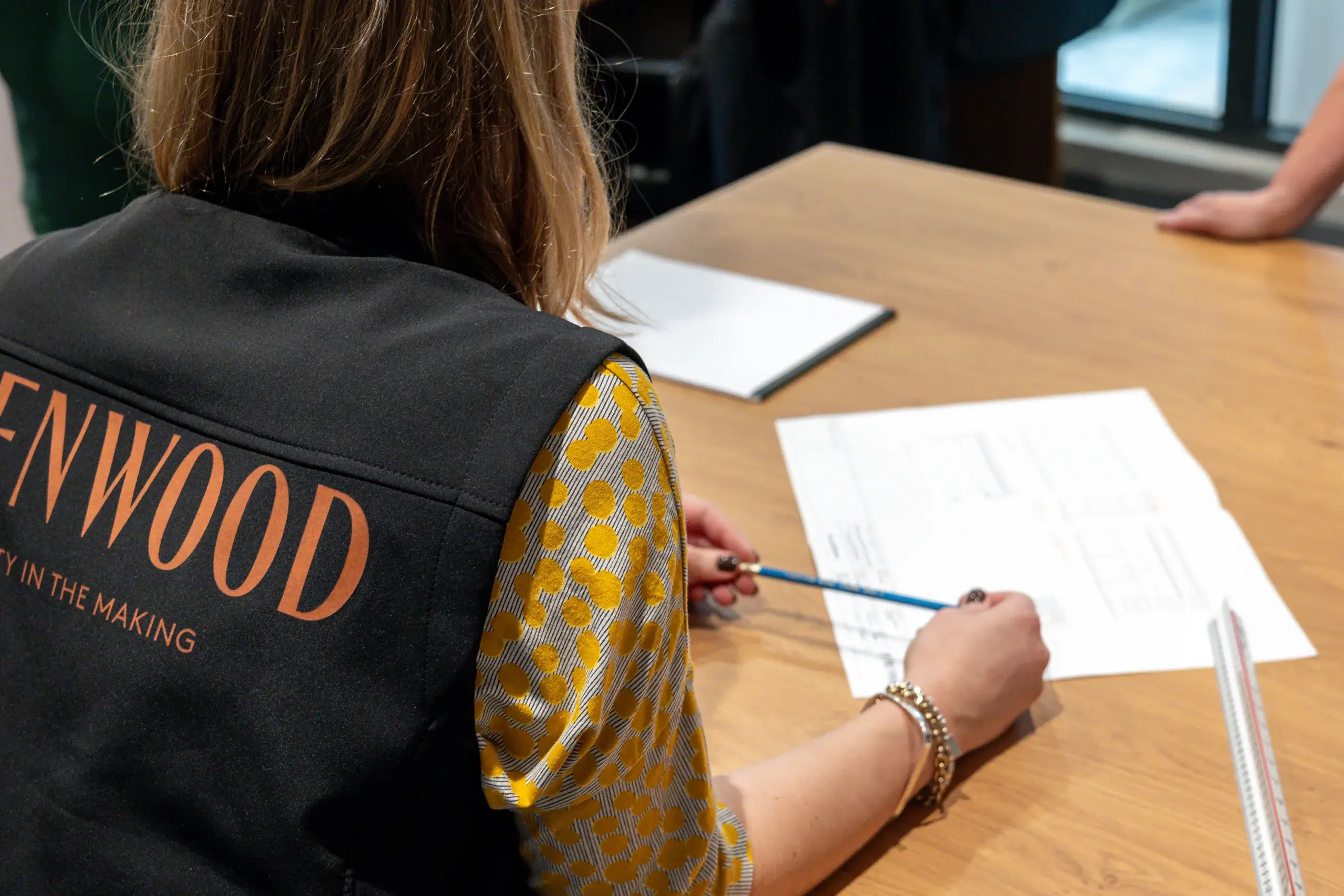 Person in a black vest with orange "WOOD" lettering leaning over a wooden table, holding a pencil and reviewing printed plans or documents.