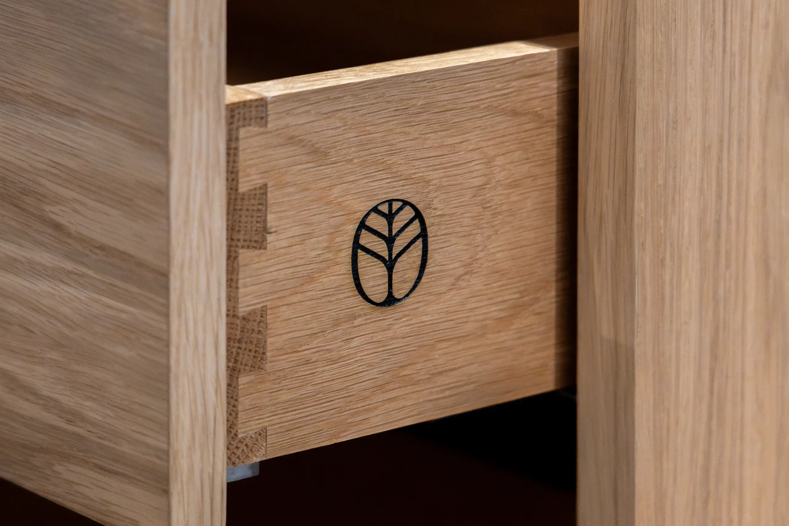 Oak drawer front with visible dovetail joints and a small black tree-like logo burned into the wood.