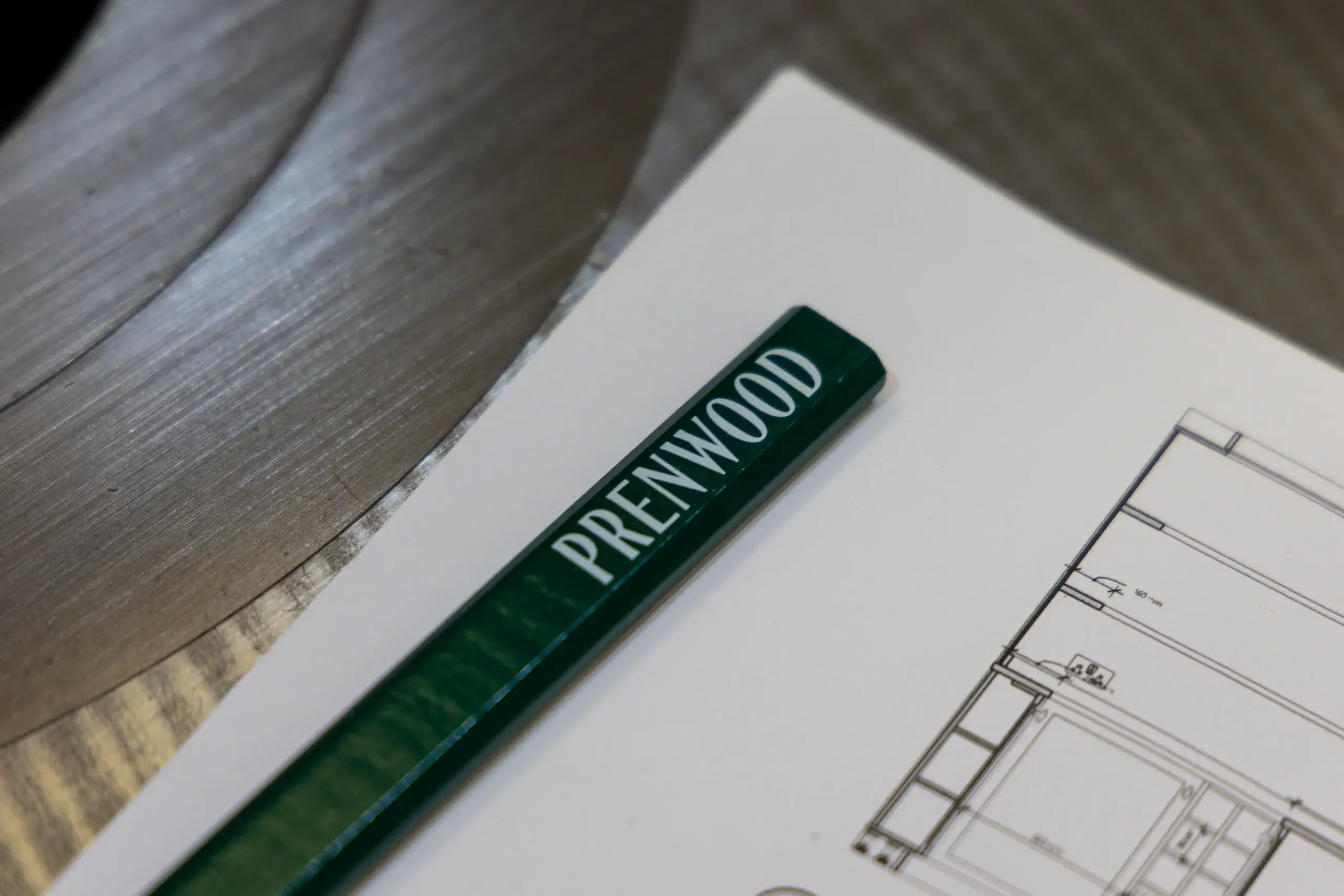 Green pencil with "PRENWOOD" printed on it resting on architectural drawings beside a metal circular saw blade.