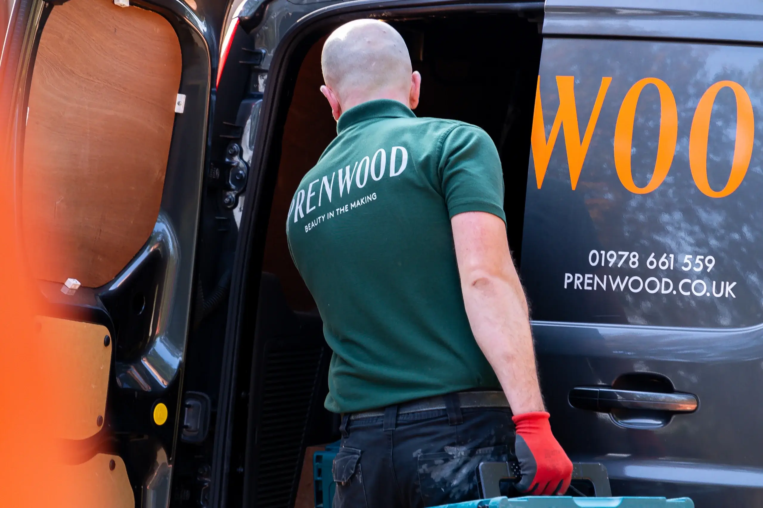 Worker in green "PRENWOOD" polo loading tools into a company van, wearing red glove and holding a blue toolbox.