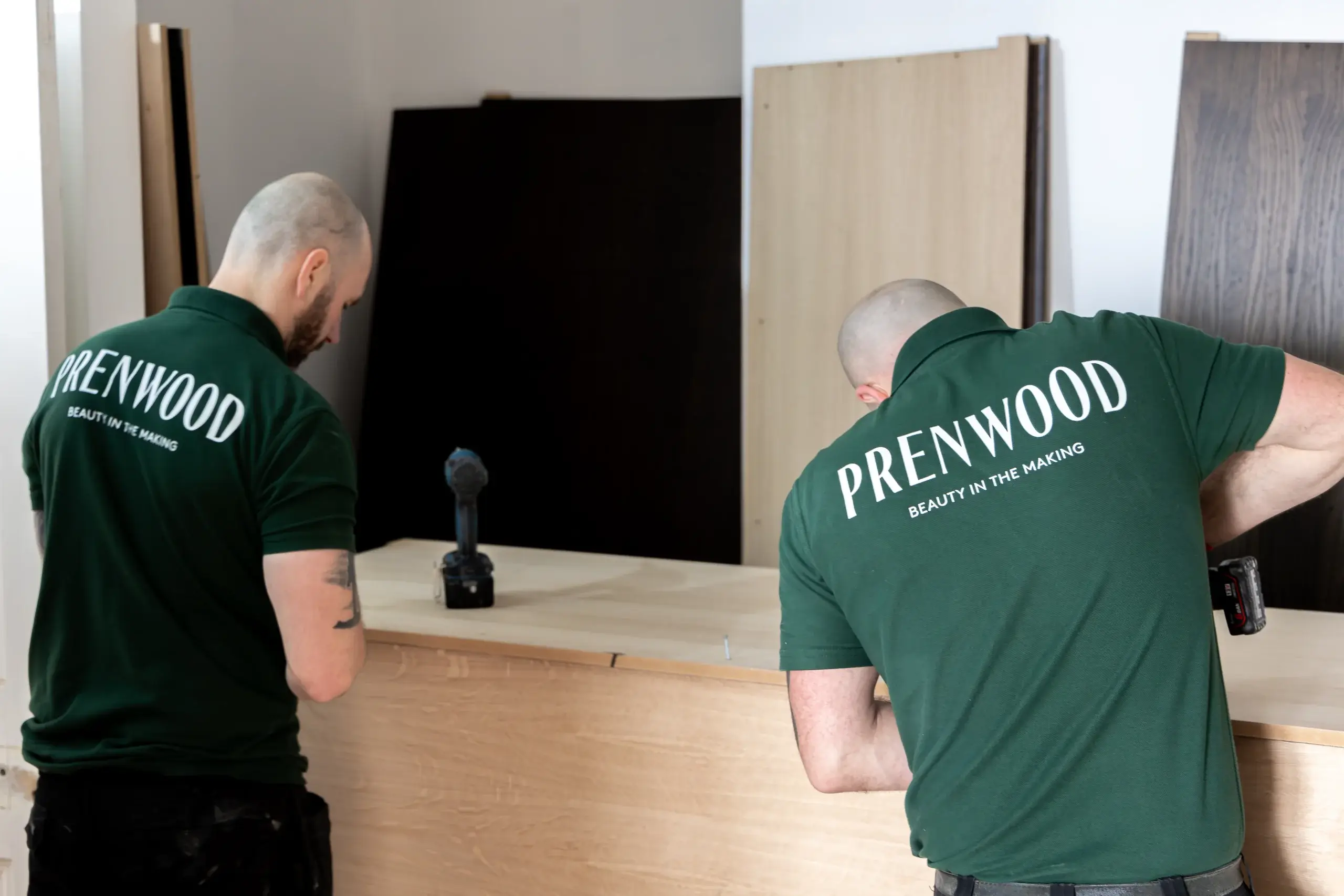 Two men in green "PRENWOOD" shirts assembling wooden panels with a cordless drill on a workbench.