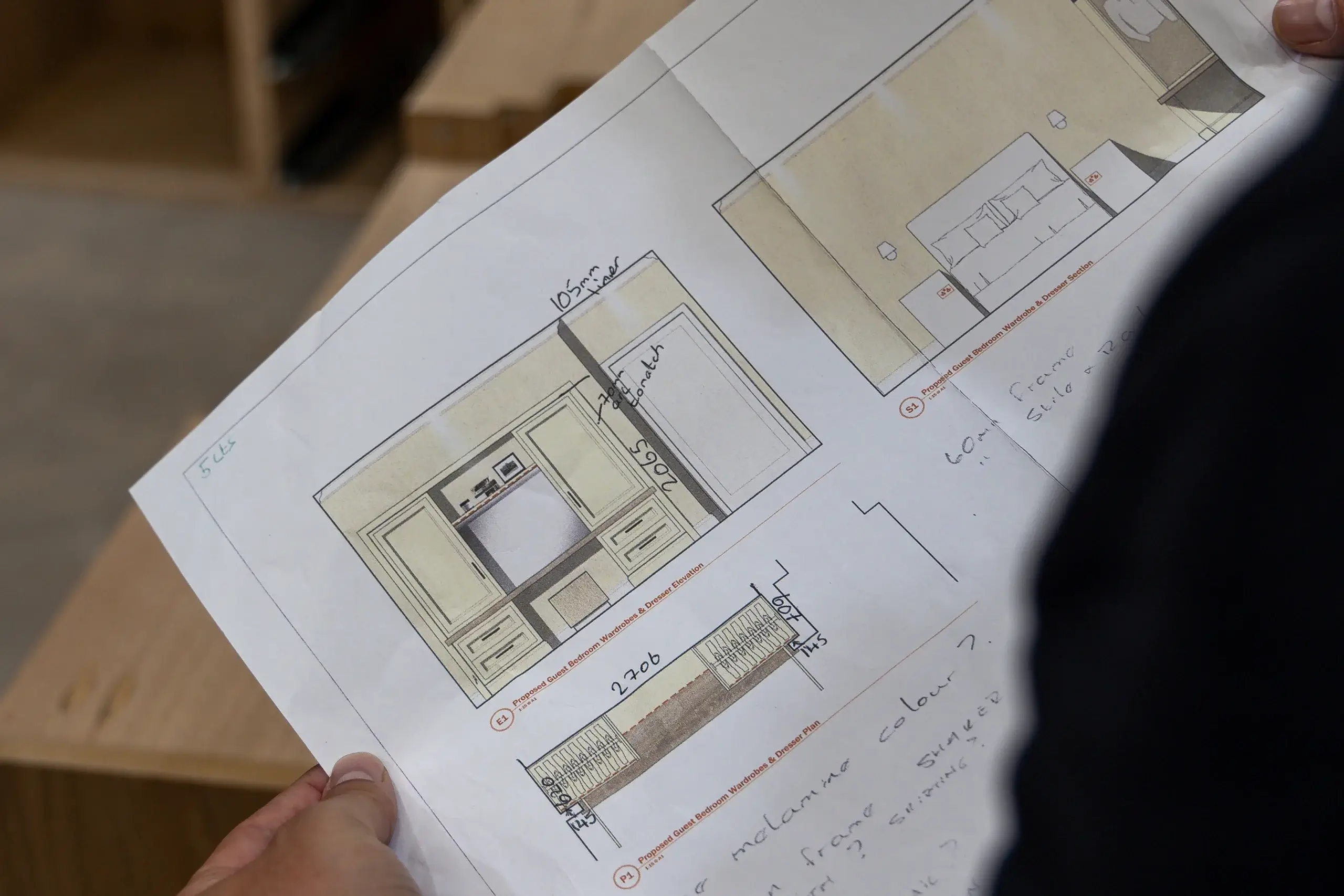 Hand holding printed furniture elevation and plan drawings with handwritten measurements and notes, showing wardrobe and drawer designs.