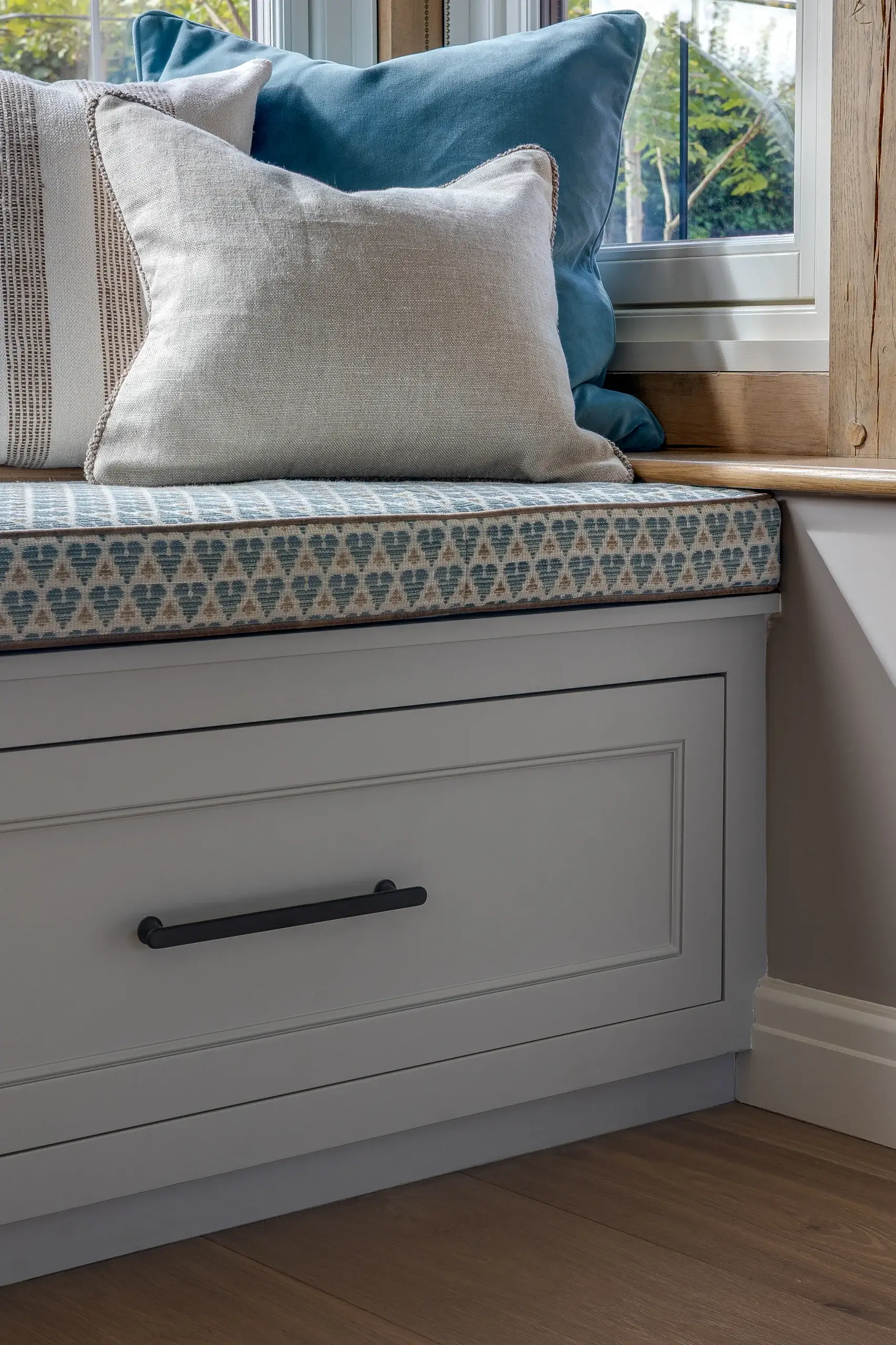 Window seat with patterned cushion, beige and teal throw pillows, and a gray built-in drawer with black handle