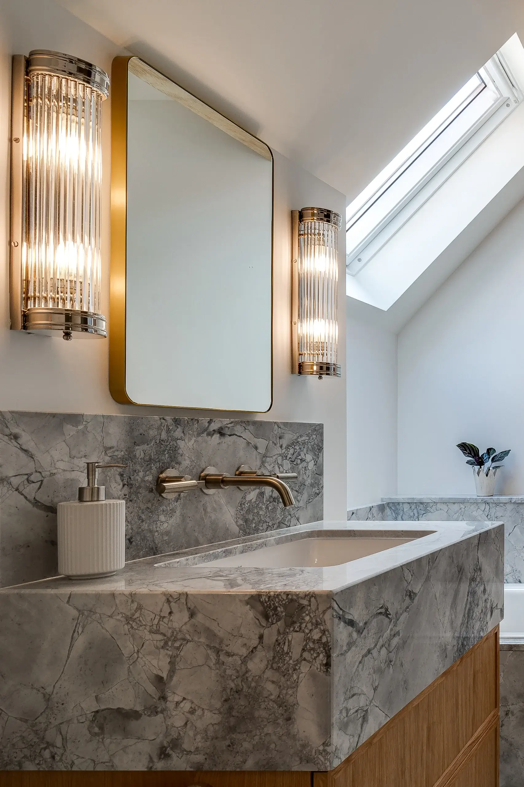 Modern bathroom sink with gray marble countertop and backsplash, wall-mounted brass faucet, gold-framed mirror and vertical ribbed sconces, skylight above.