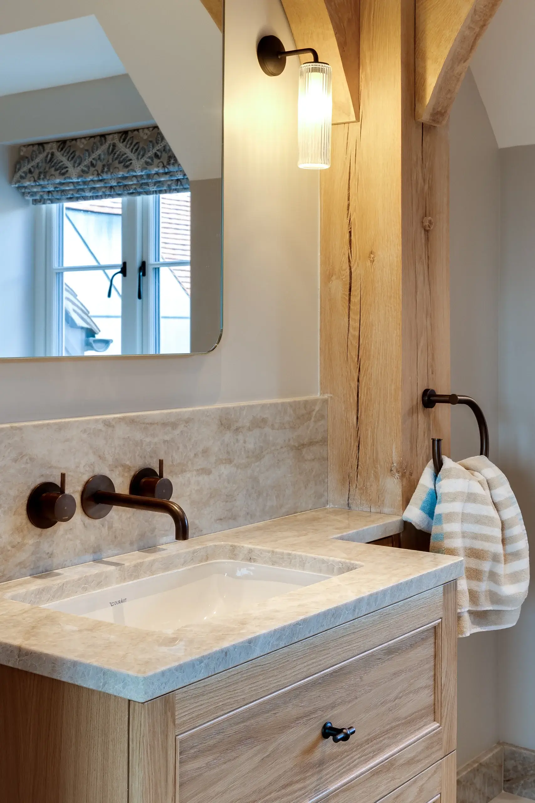 Bathroom vanity with marble countertop, undermount sink, wall-mounted bronze faucet, wood cabinetry and striped hand towel on ring
