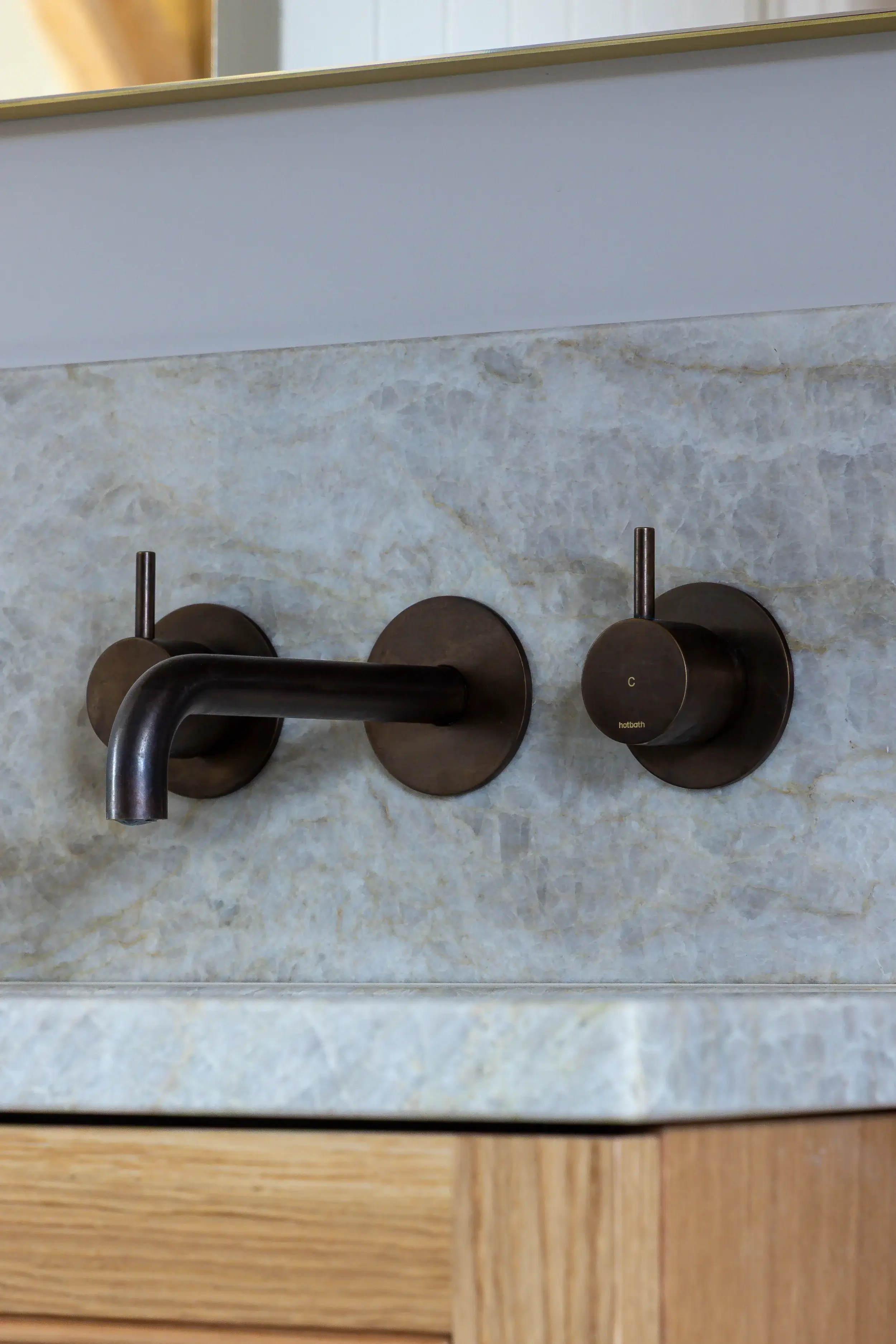 Close-up of a wall-mounted bronze bathroom tap and dual handles set against a soft-toned stone splashback, with oak cabinetry beneath.