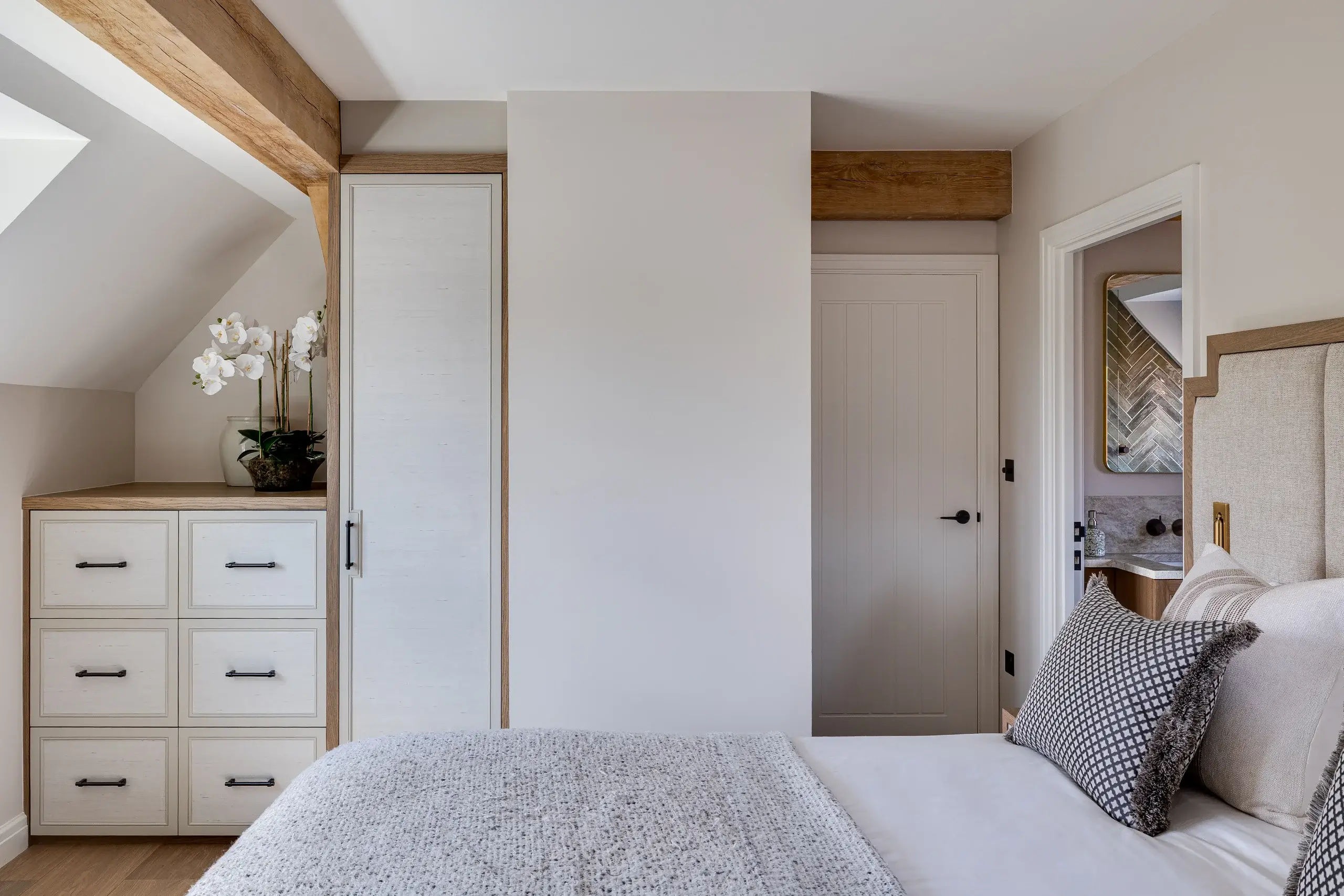 Neutral-toned bedroom corner with built-in white drawers and wardrobe, wooden beams, white orchid on top, and a bed with textured gray pillows.