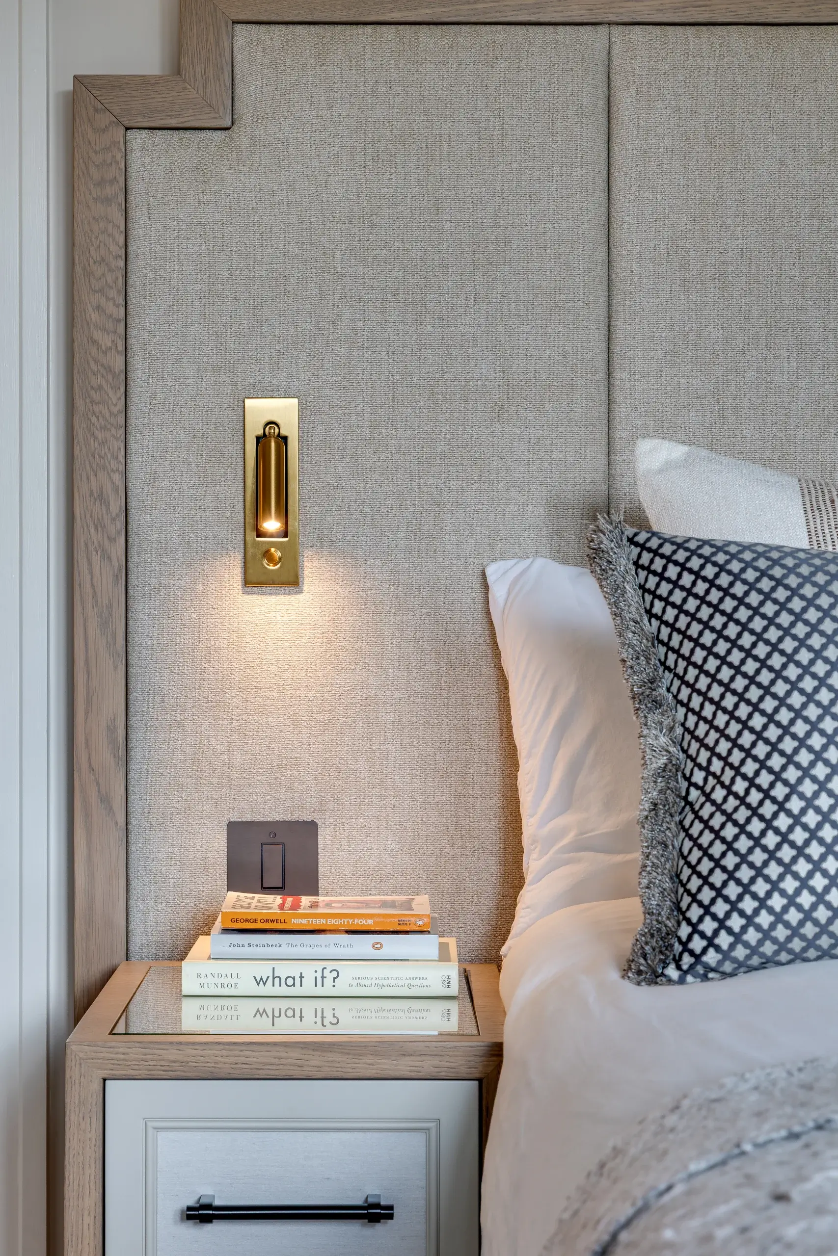 Bedside vignette with beige upholstered headboard, brass recessed reading light, stack of books on a mirrored nightstand and patterned throw pillow.