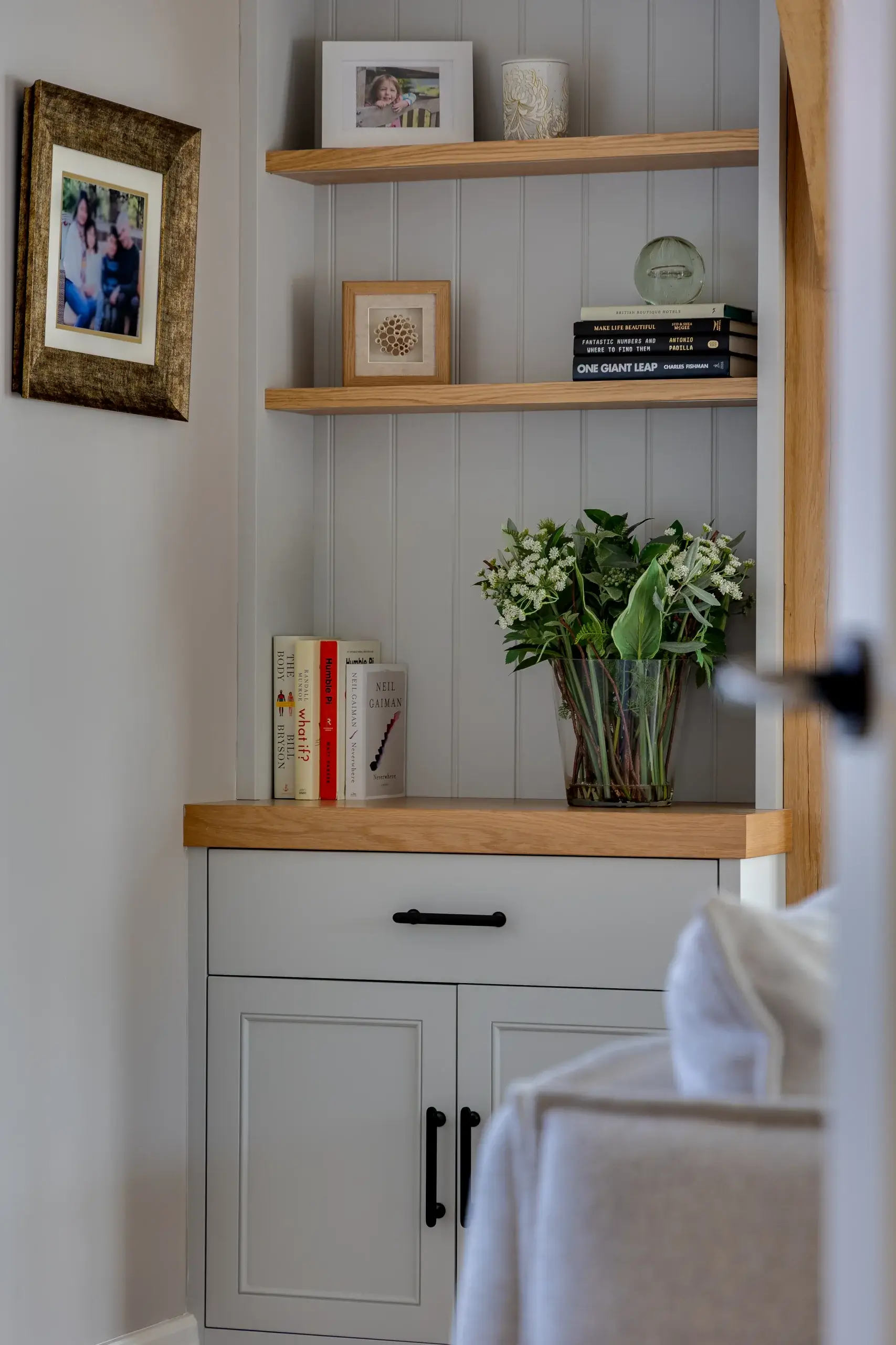 Narrow built-in shelving with wood trim holding framed photos, books and a glass vase of green-and-white flowers above a gray cabinet