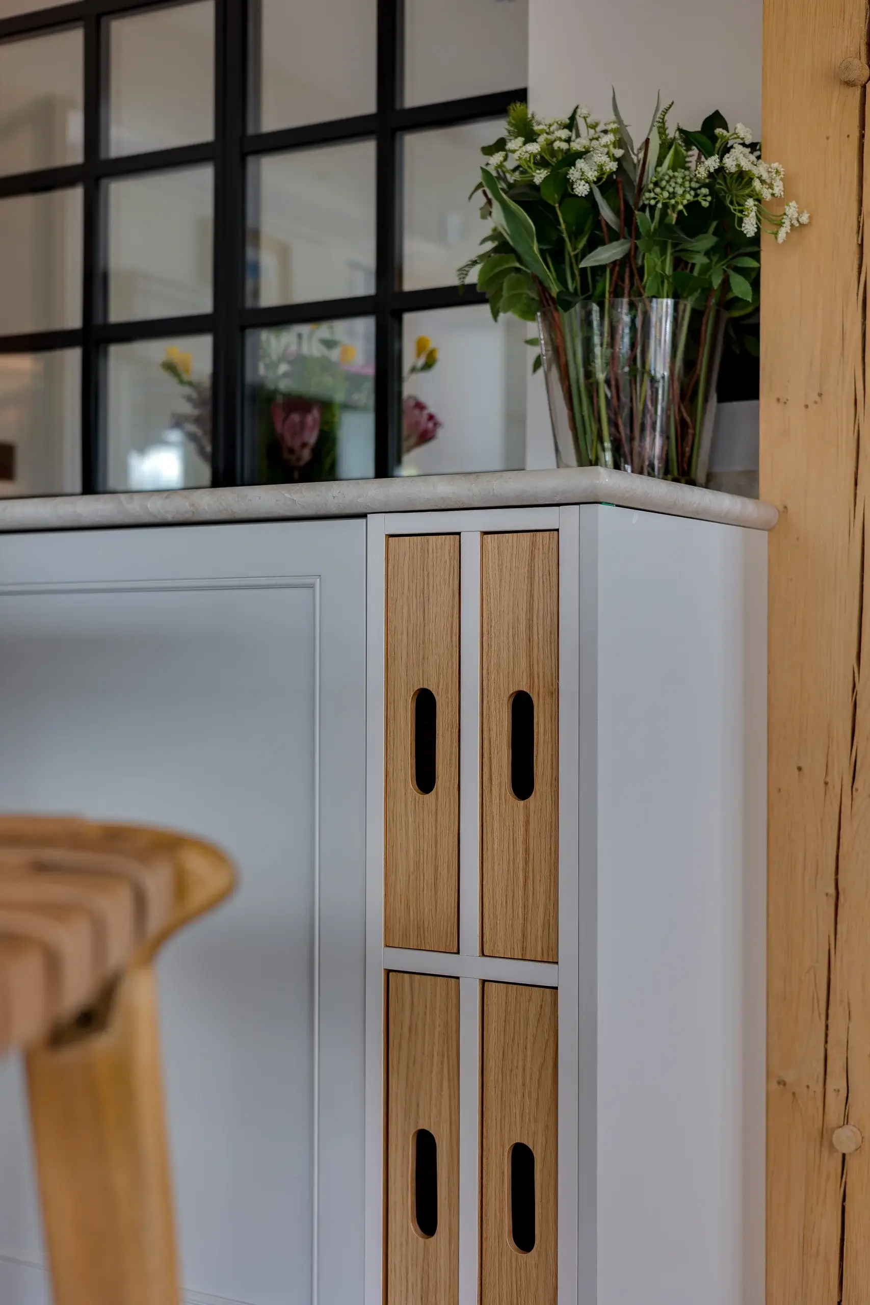 Kitchen island corner with built-in oak storage panels, marble countertop, glass vase of green and white flowers, and stool edge visible