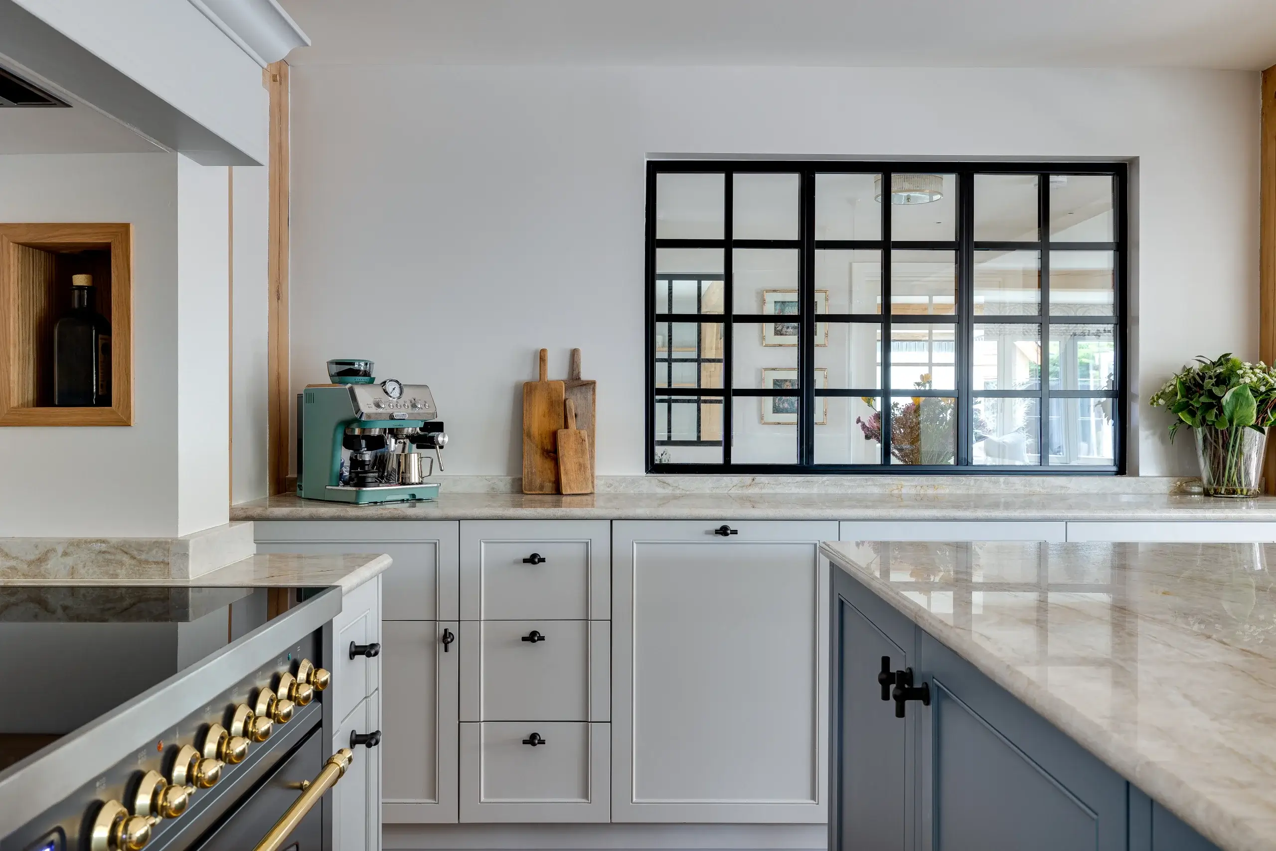 Bright modern kitchen countertop with marble surfaces, gray cabinets, teal espresso machine, wooden cutting boards and black-framed pass-through window reflecting adjacent room.