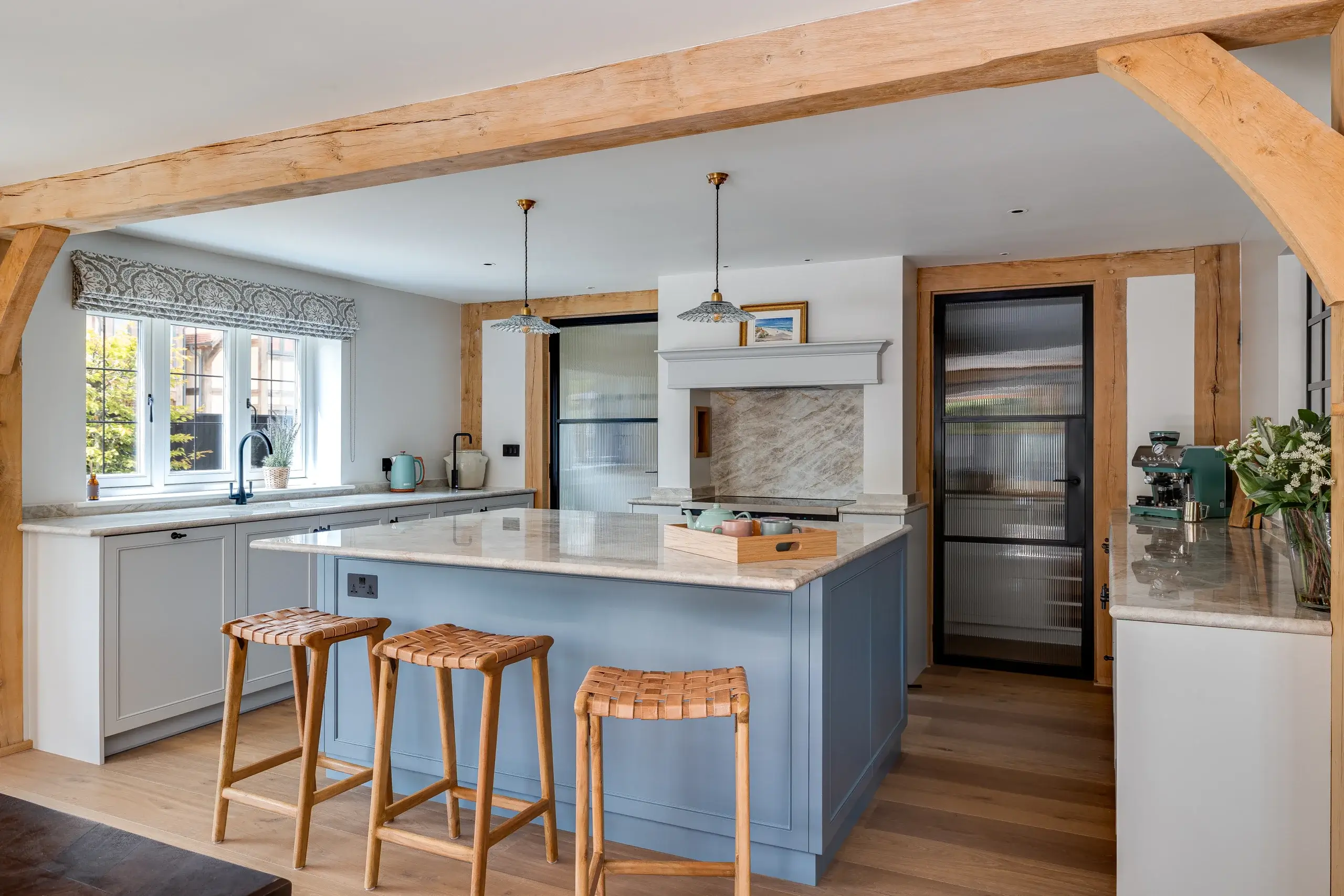 Bright farmhouse kitchen with a blue central island topped with marble, three woven wooden stools, exposed timber beams, and pendant lights.