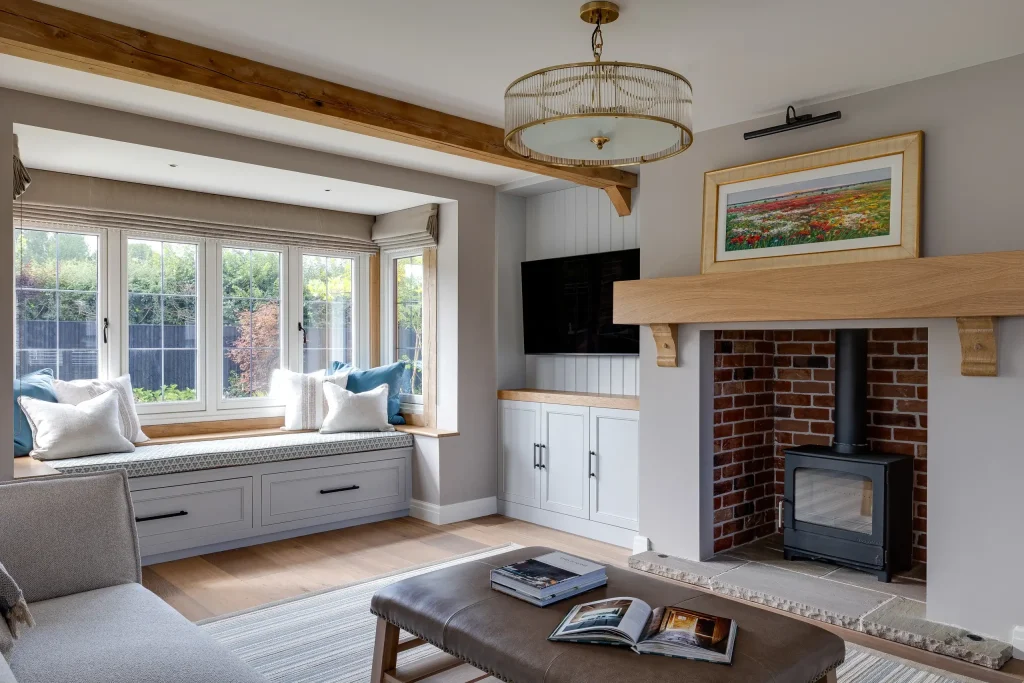 Cozy living room with window seat and cushions, wood-burning stove in brick fireplace, neutral decor and oak beams