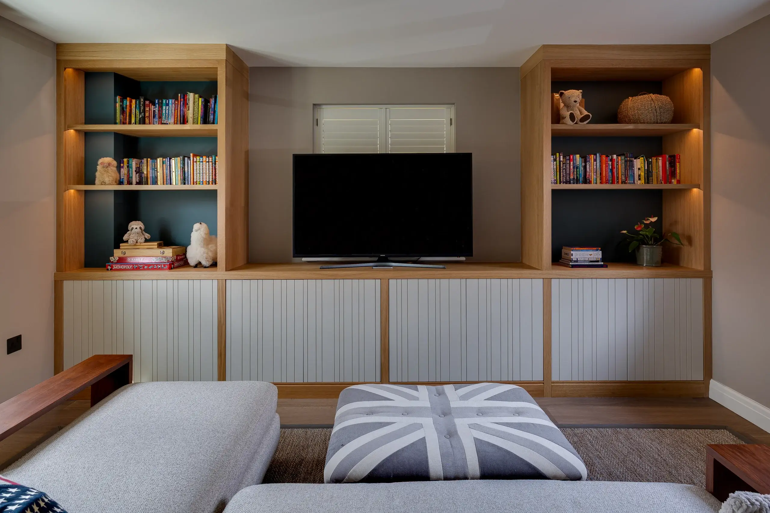 Built-in wooden media unit with central flat-screen TV, flanked by lit bookshelves holding books, toys and plants, and gray paneled cabinets below.