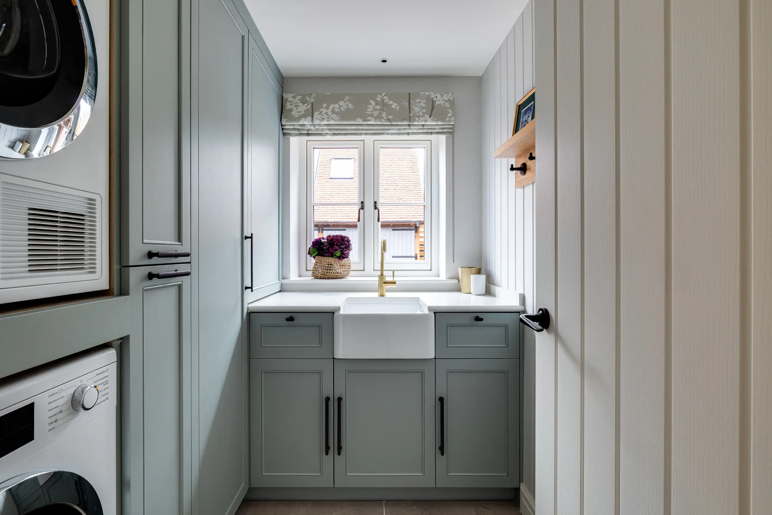 A stylish utility room with sage green cabinetry, a farmhouse sink, and integrated laundry appliances, brightened by a patterned roman blind and natural light.