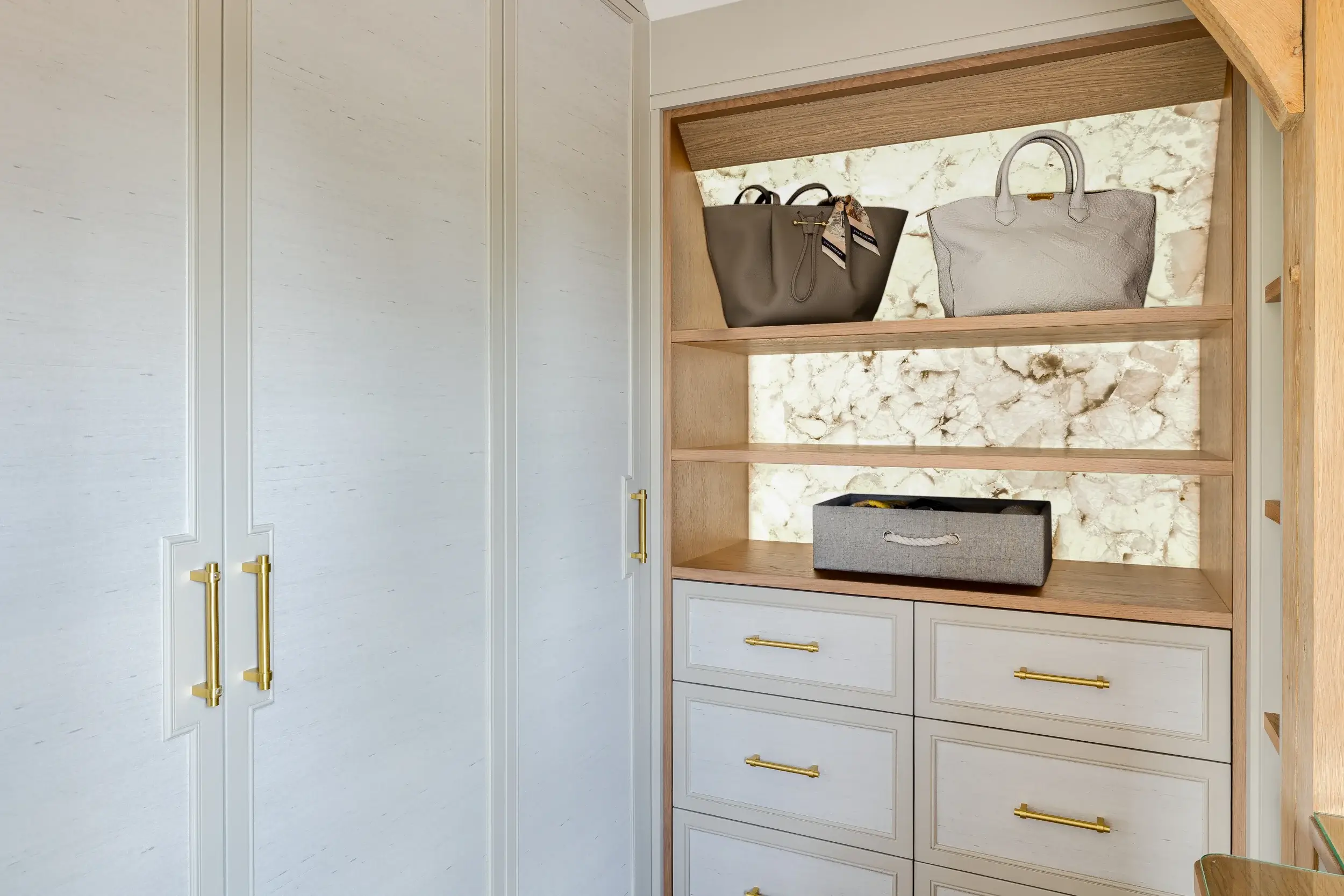 A bespoke wardrobe with pale painted doors and brushed gold handles, complemented by open oak shelving with a backlit stone feature, displaying handbags.