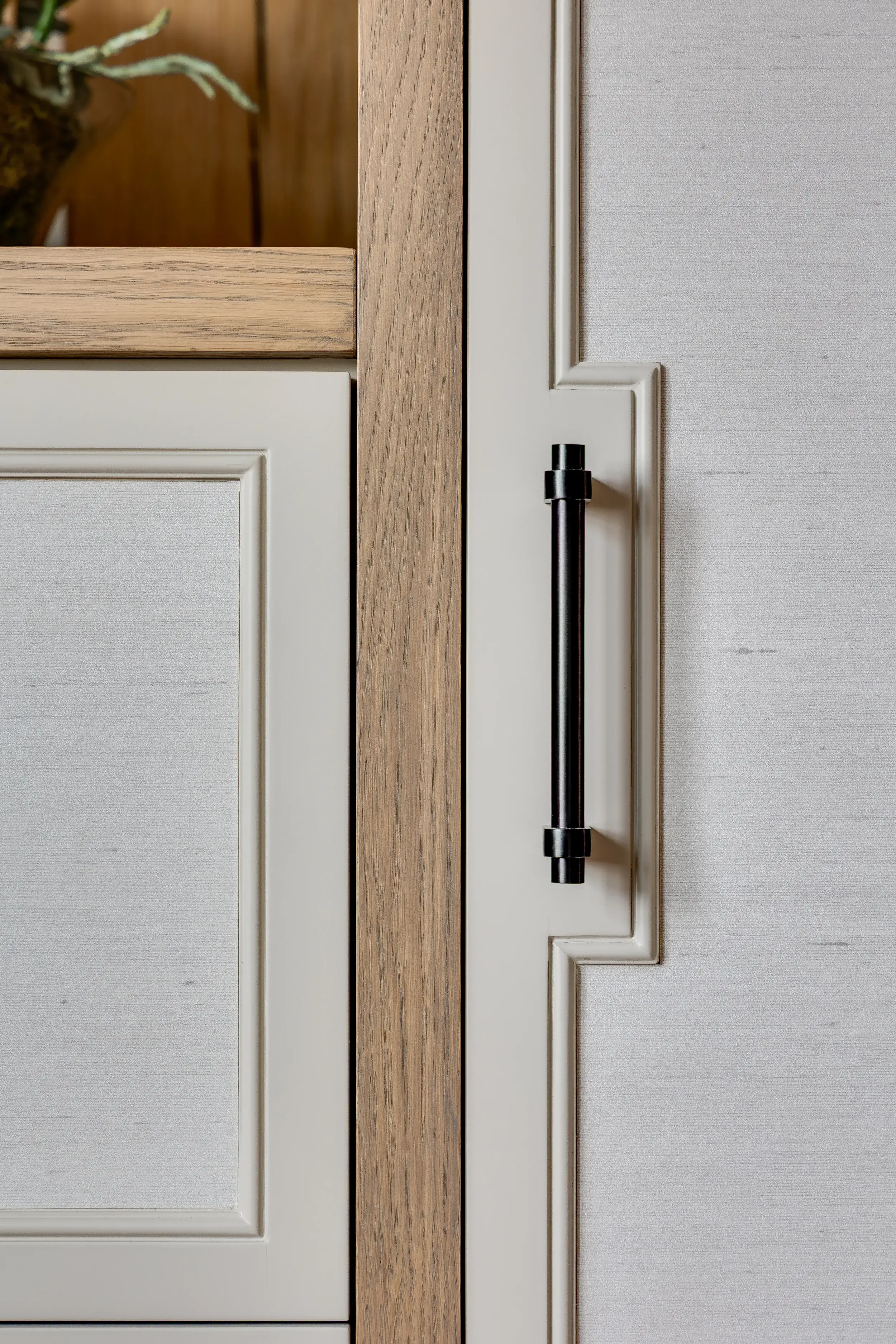 A close-up of bespoke wardrobe joinery with pale painted panels, oak trim, and a sleek black handle, showcasing craftsmanship and texture.