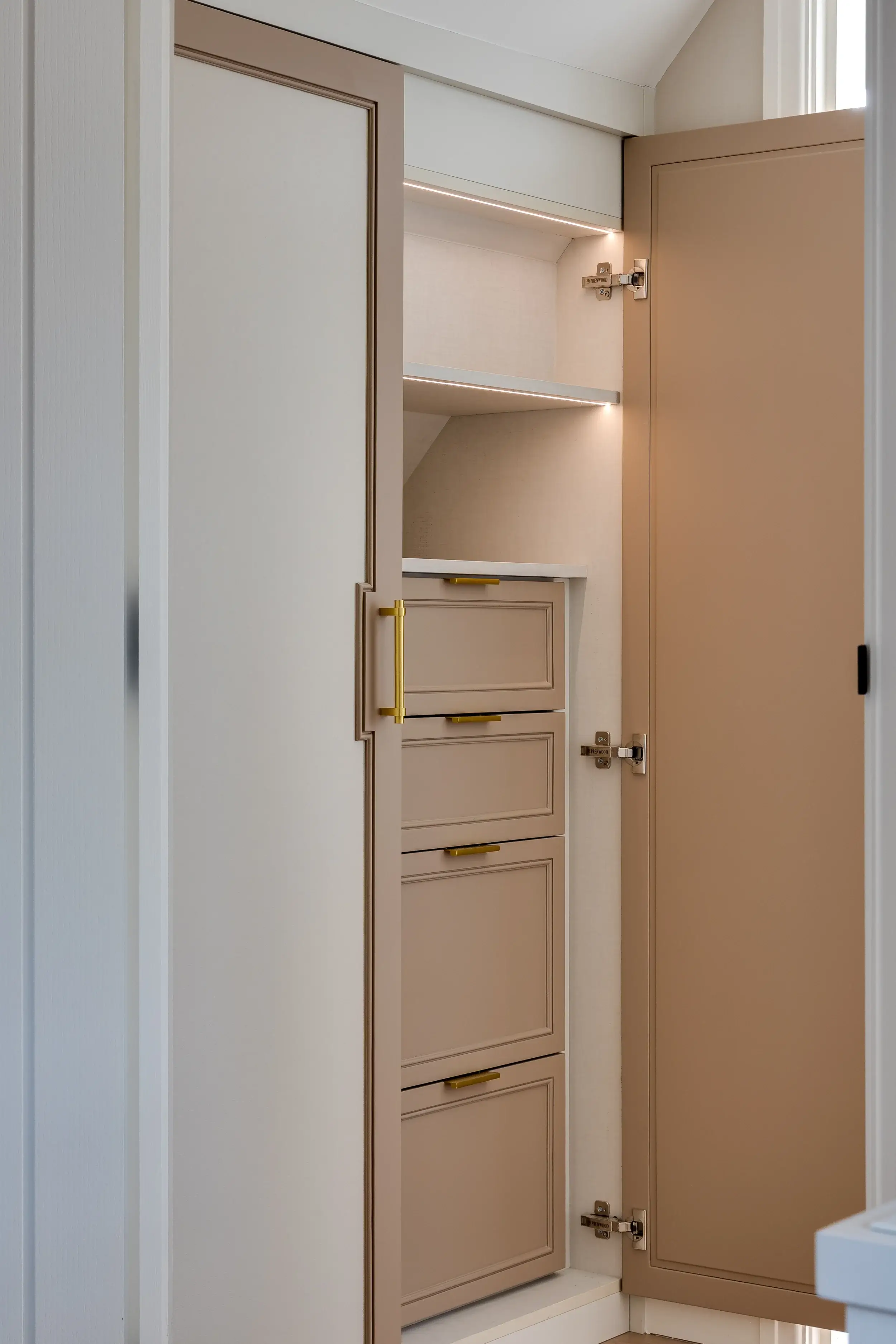 A bespoke fitted wardrobe in a soft beige finish, featuring gold handles, open shelving with integrated lighting, and neatly hidden drawers for elegant storage.
