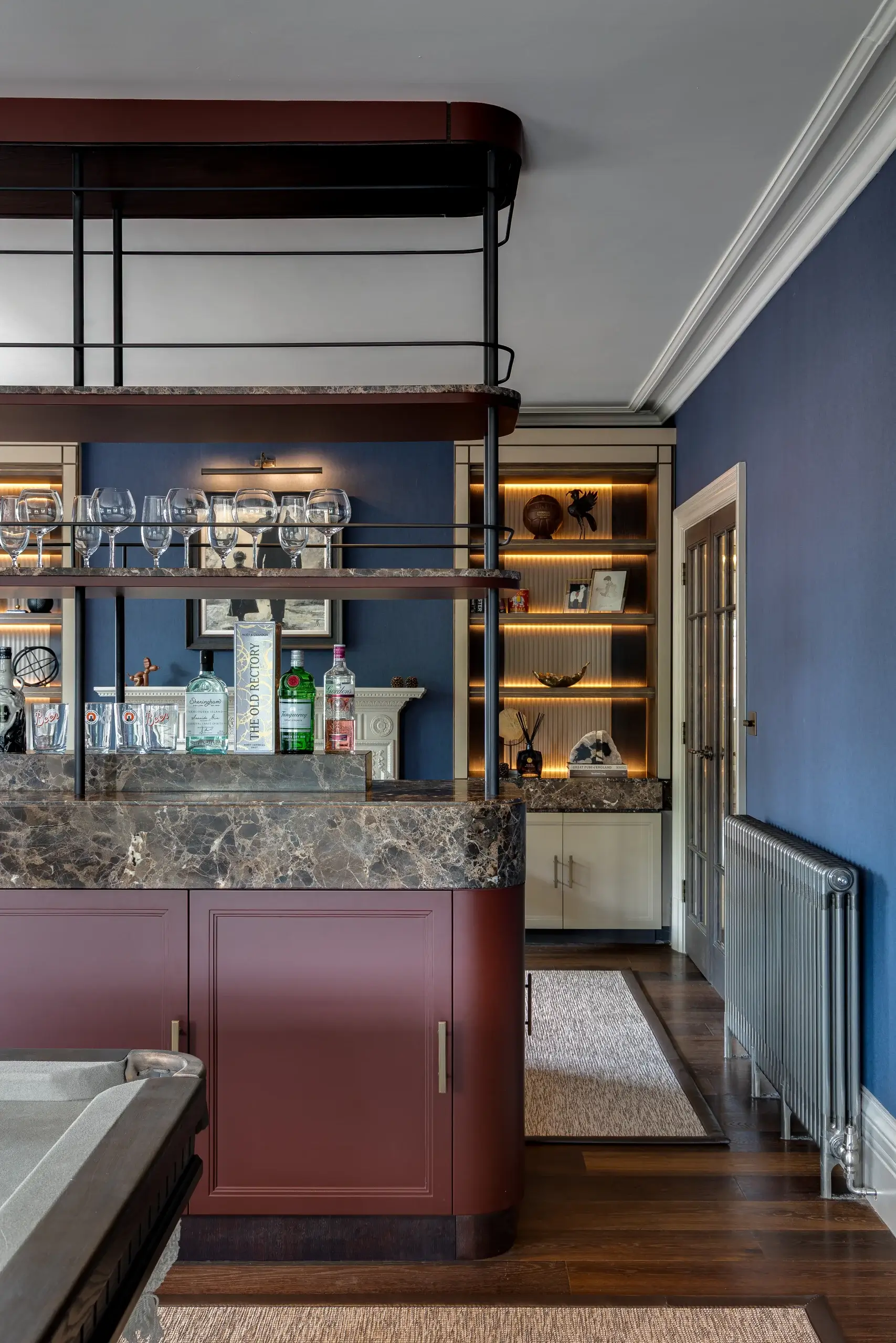 Home bar with marble countertop, burgundy cabinets, glassware on metal shelving and illuminated built-in shelving against blue walls.