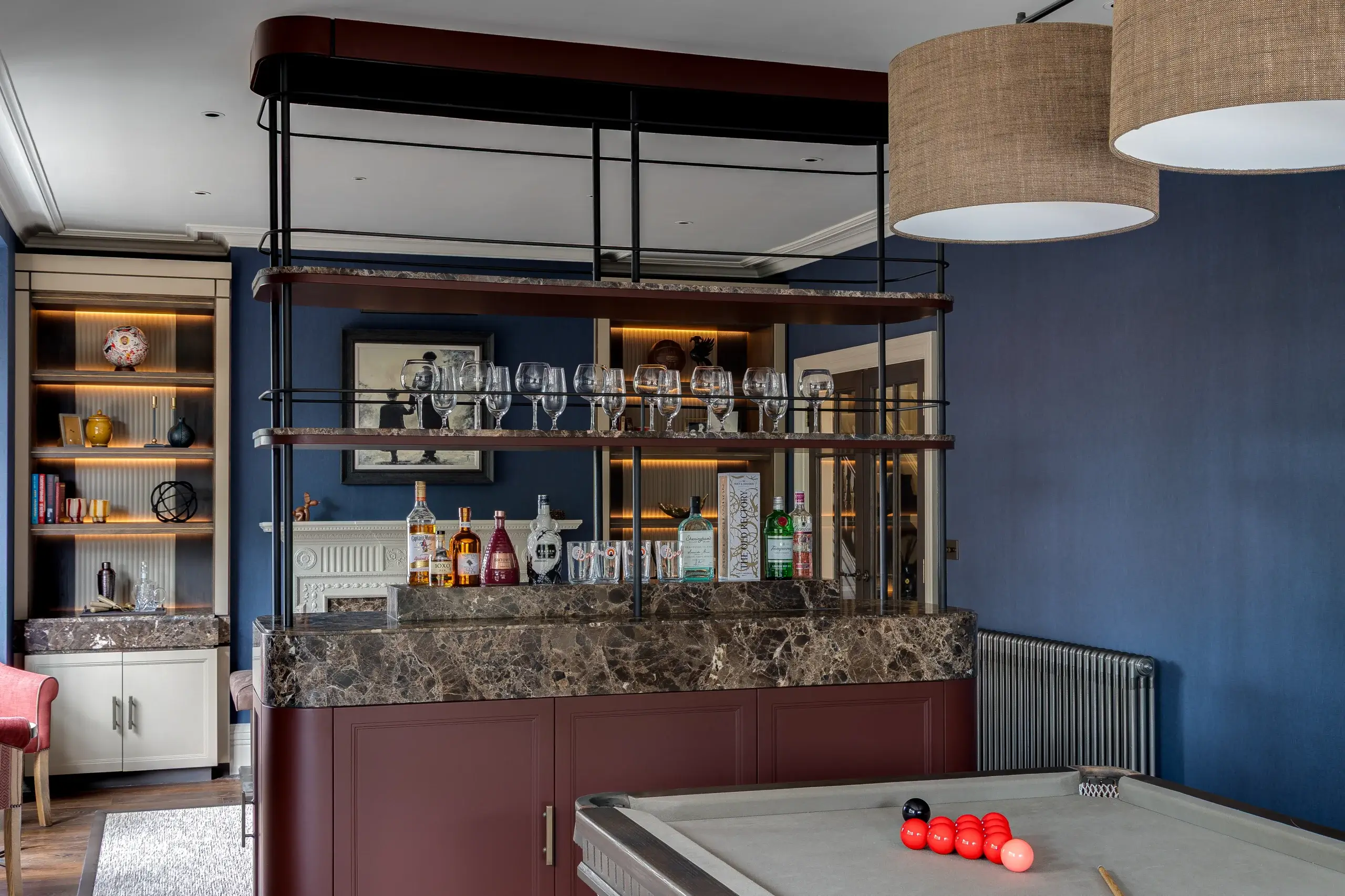 Home bar with marble counter, shelves of liquor and glasses, blue walls, and a pool table with red balls in foreground.