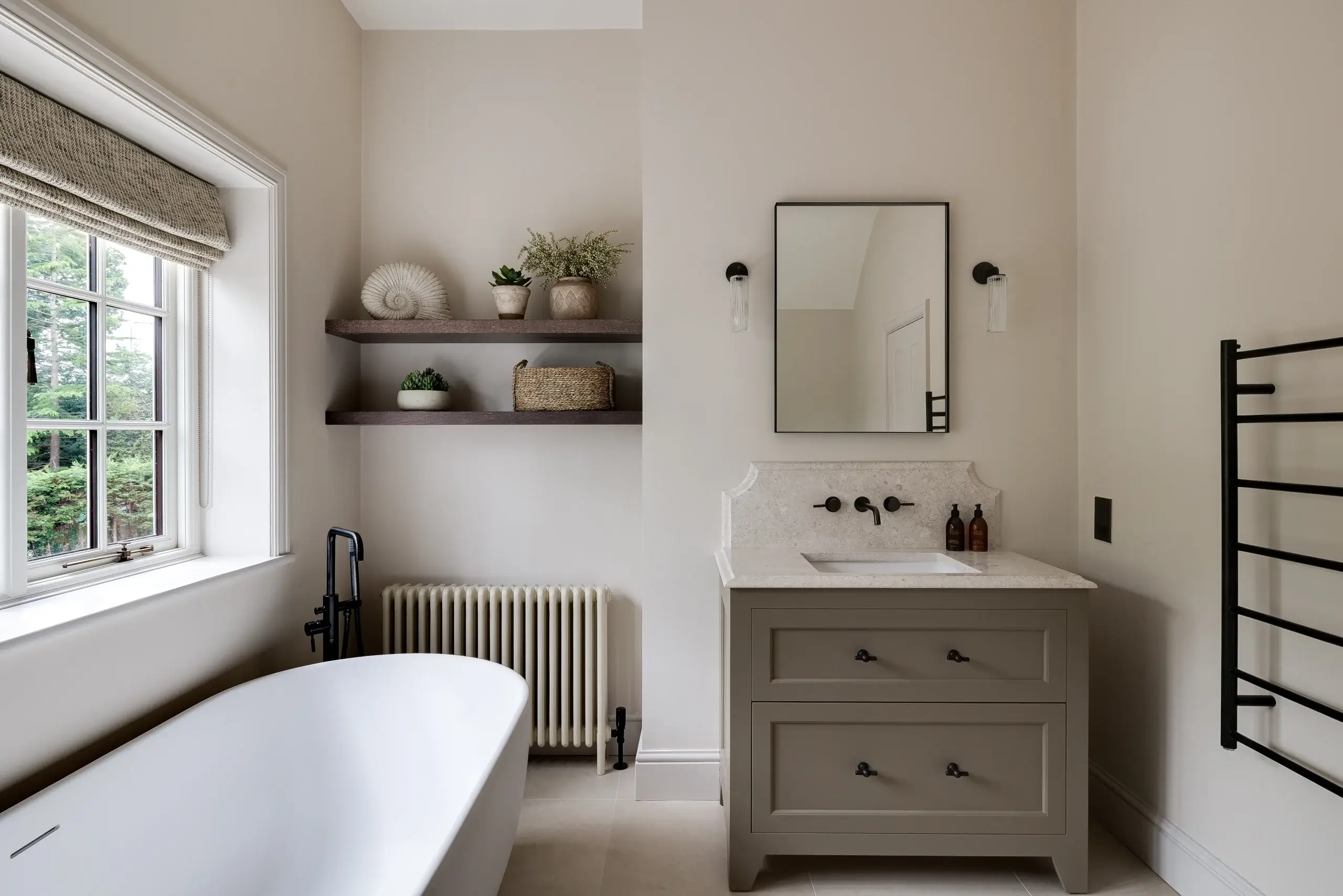 Neutral-toned bathroom with freestanding tub, beige vanity with mirror, wall shelves with plants and basket, window light.