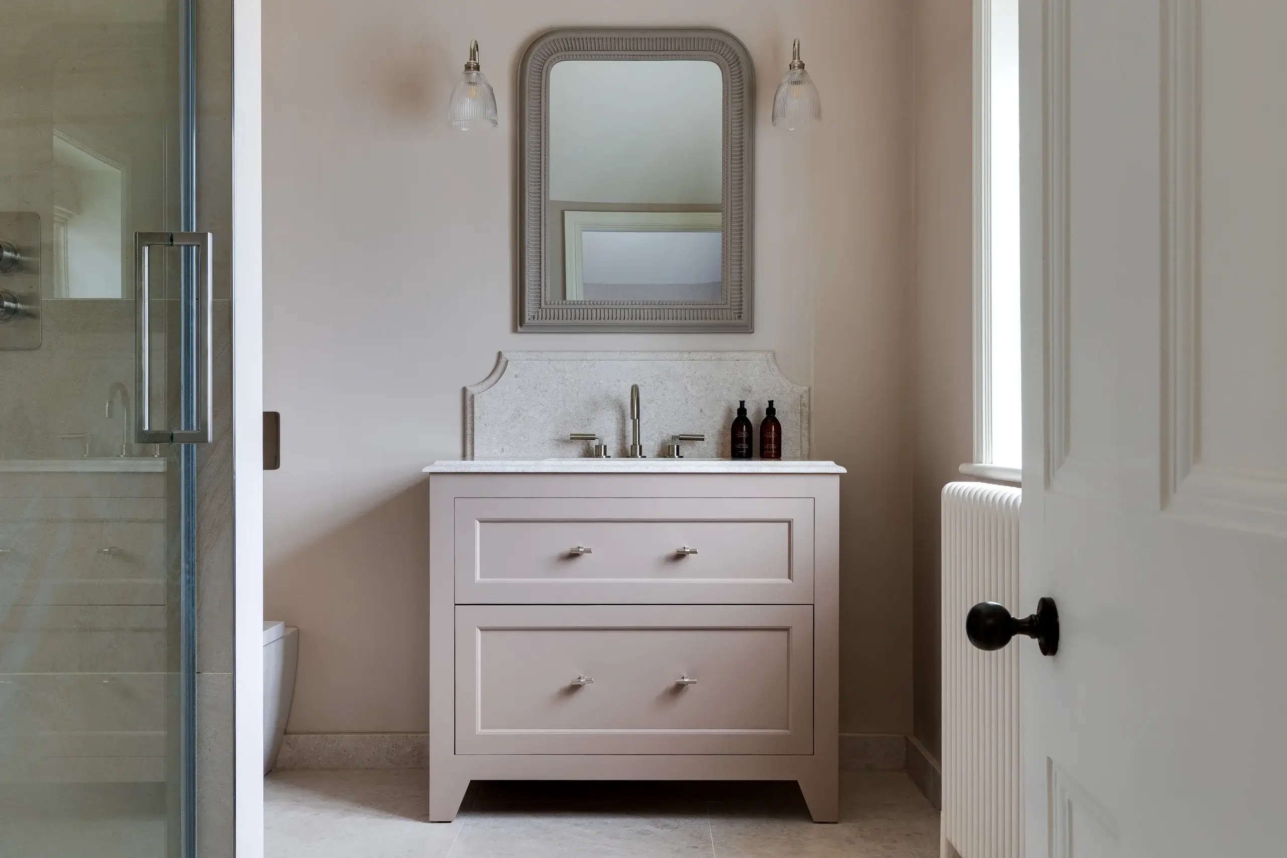 Muted pink bathroom vanity with marble countertop, centered faucet, two brown soap bottles, framed mirror and wall sconces above.
