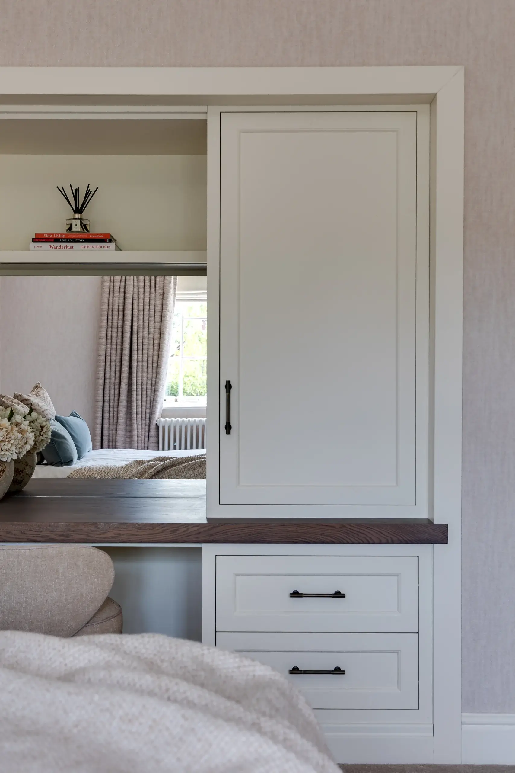 Built-in white cabinetry with dark wood countertop, drawers with black handles, and a small shelf holding books and a reed diffuser, bedroom seen through opening.