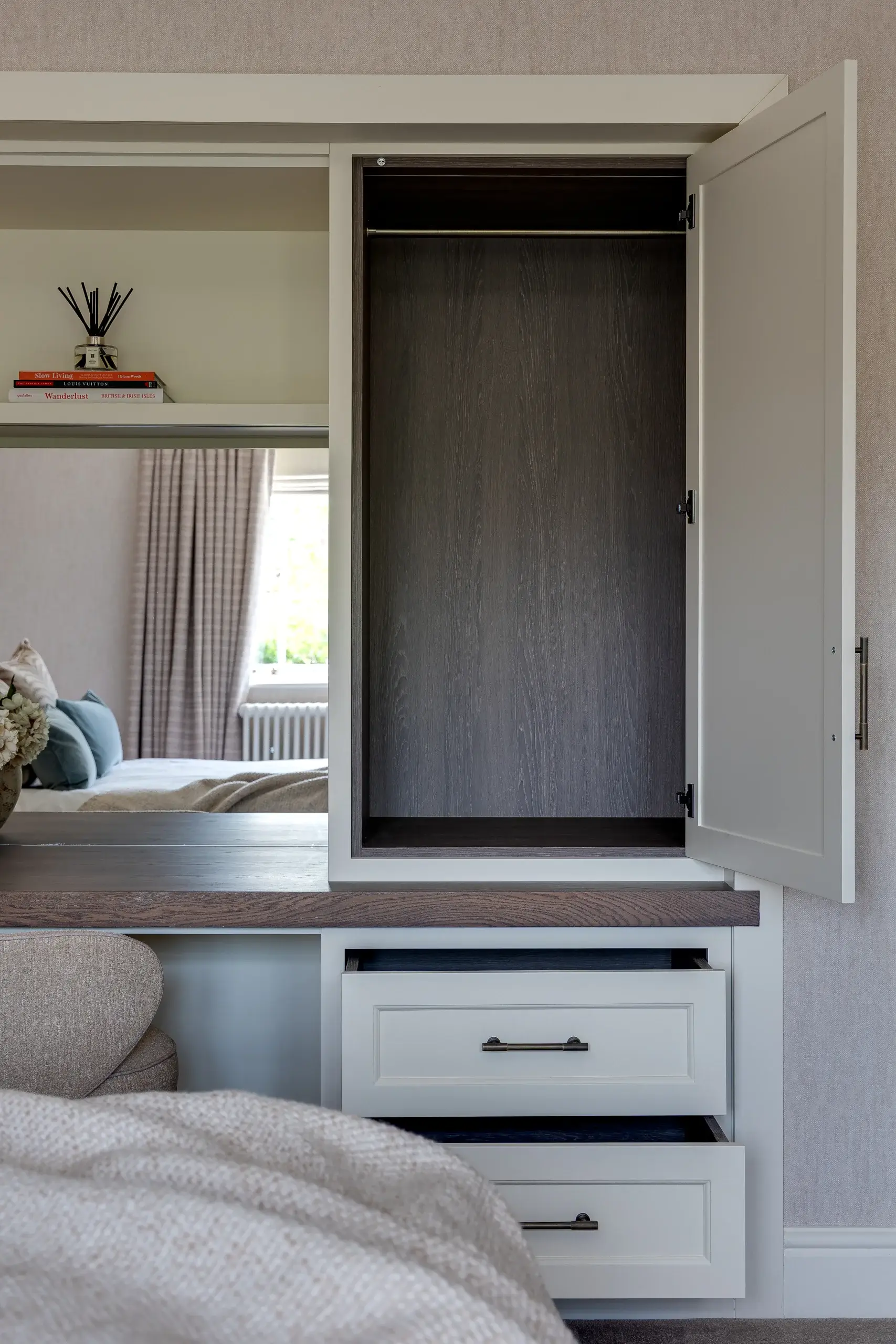 Built-in bedroom cabinet with an open tall cupboard, two partially open white drawers, wood countertop and a mirror reflecting a bed and window.