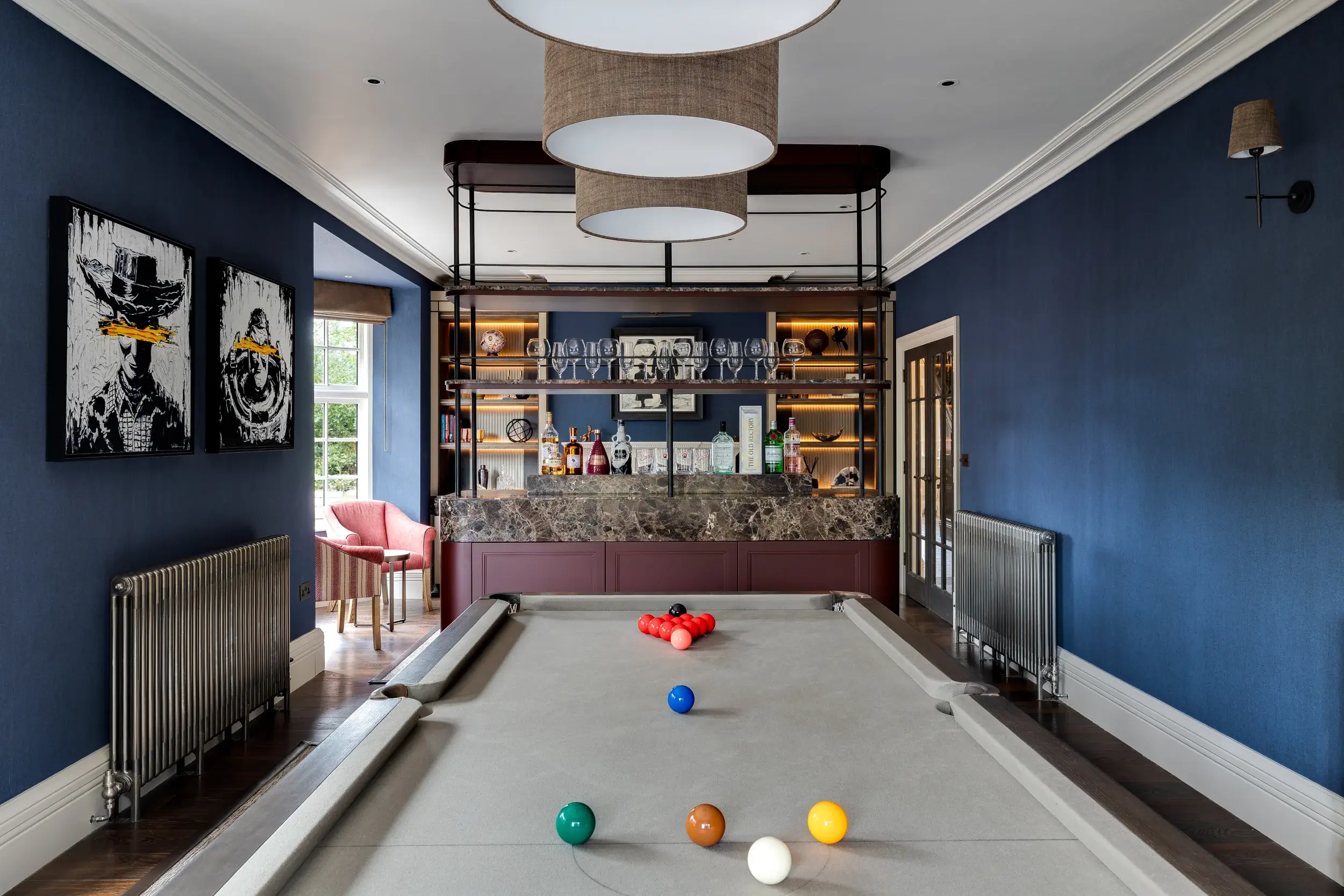 A stylish home bar and games room with deep blue walls, a grey pool table set for a game, and a marble-topped bar with illuminated shelving stocked with glassware and spirits.